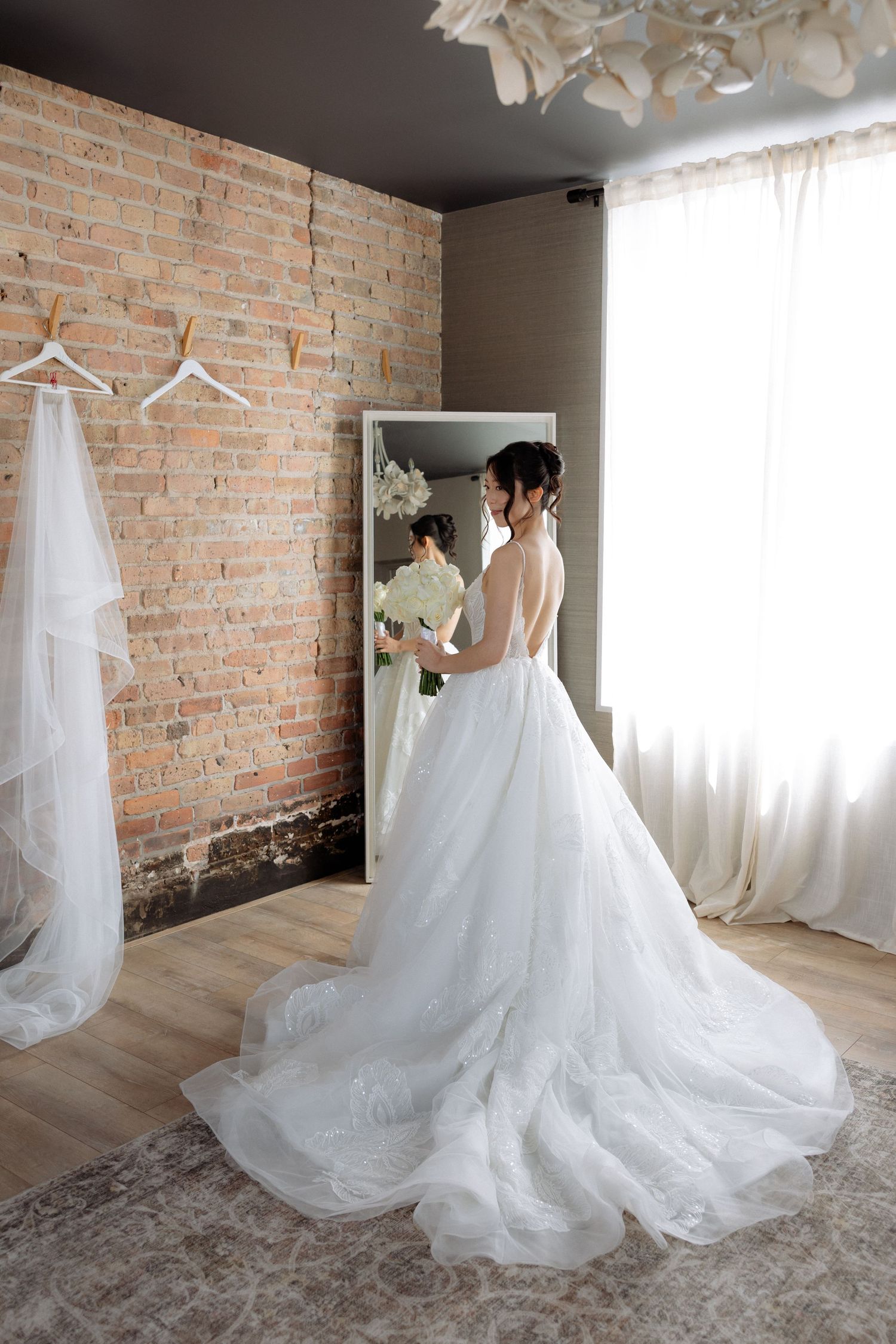 Bride in stunning backless ball gown with long train stands before mirror in rustic brick-walled room.