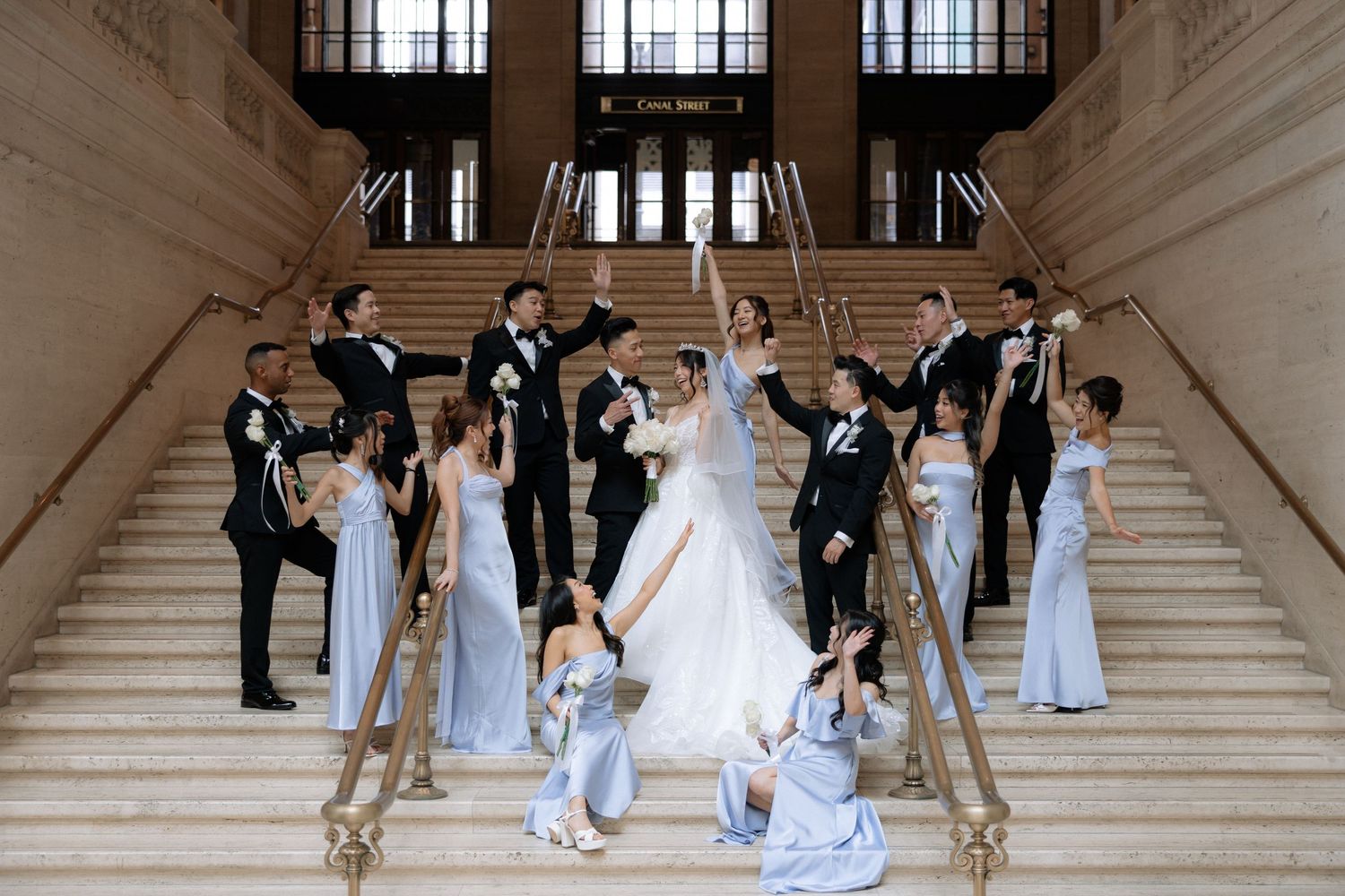 Wedding party celebrates on grand staircase, bridesmaids in blue gowns and groomsmen in black tuxedos.