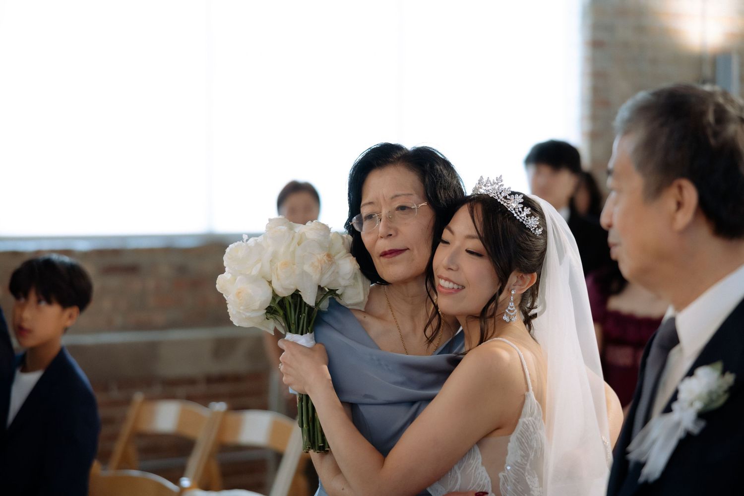Bride in tiara hugging her mother while holding white rose bouquet during emotional wedding ceremony.