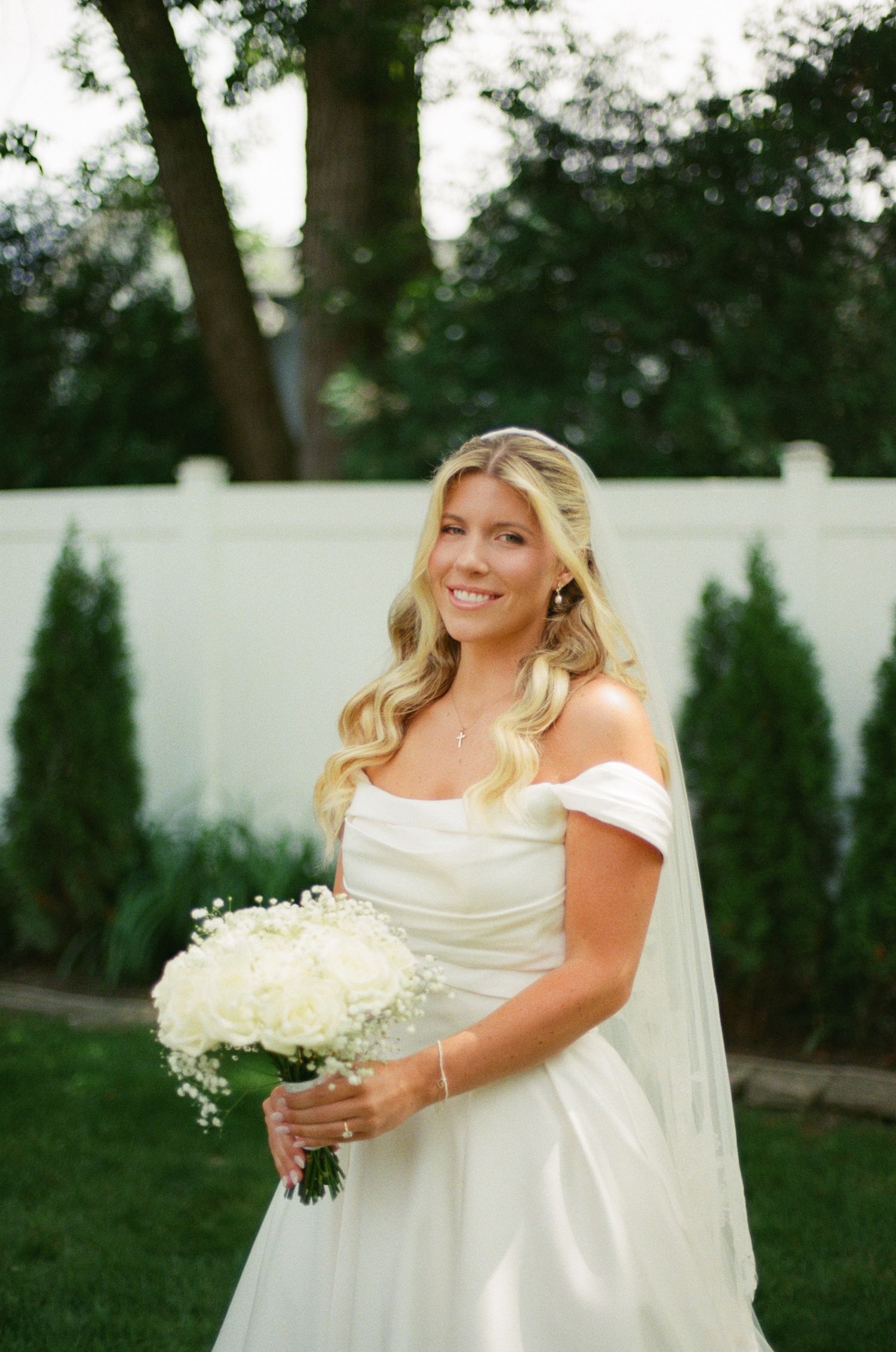 Smiling bride in off-shoulder white gown holding white rose bouquet, standing outdoors near evergreen shrubs and white fence.