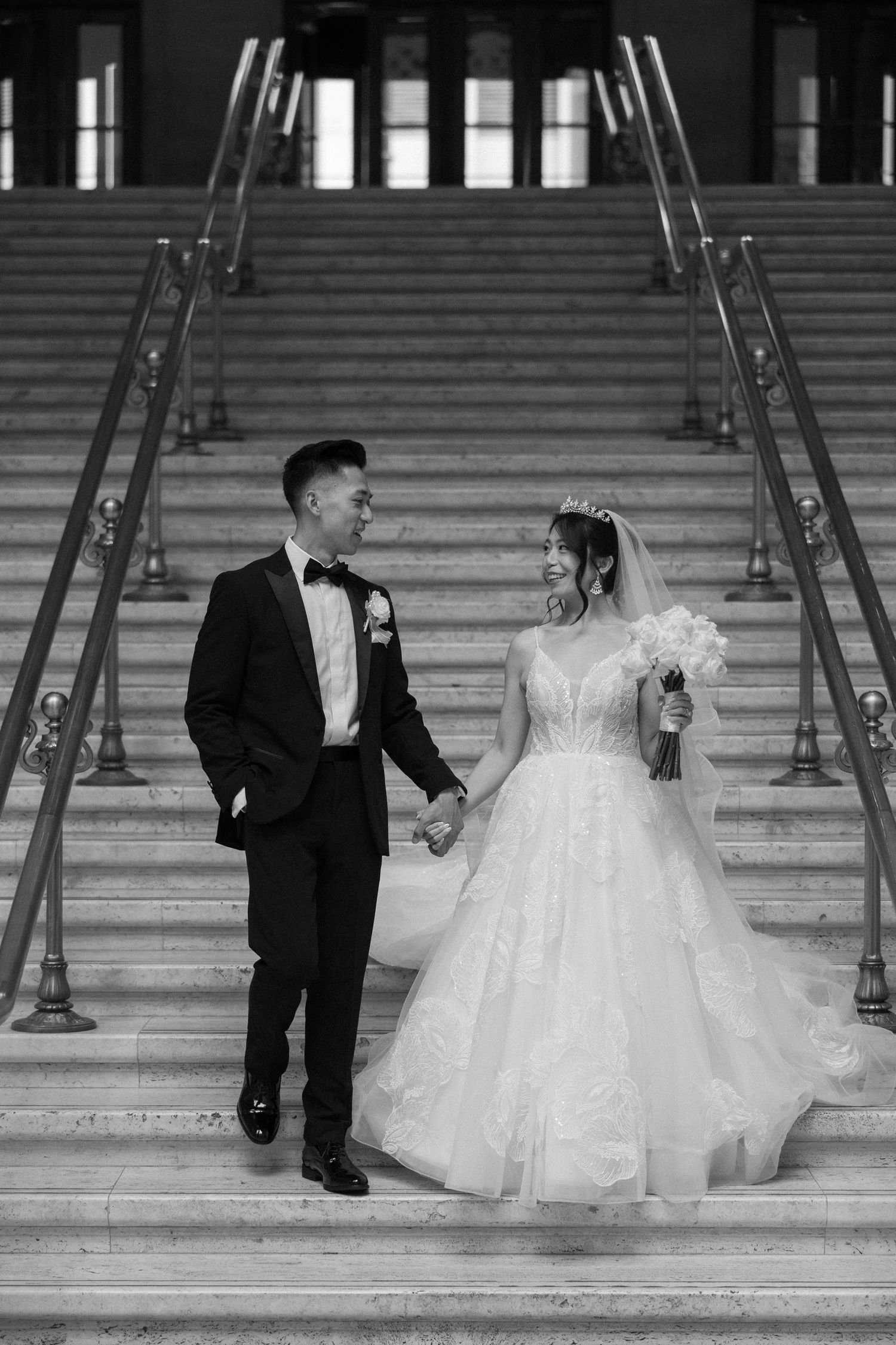 Black and white photo of bride and groom holding hands on grand staircase at wedding venue.