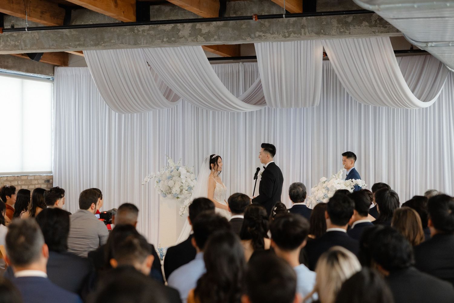 Elegant indoor wedding ceremony with white draping, floral arrangements, and guests seated as couple exchanges vows.