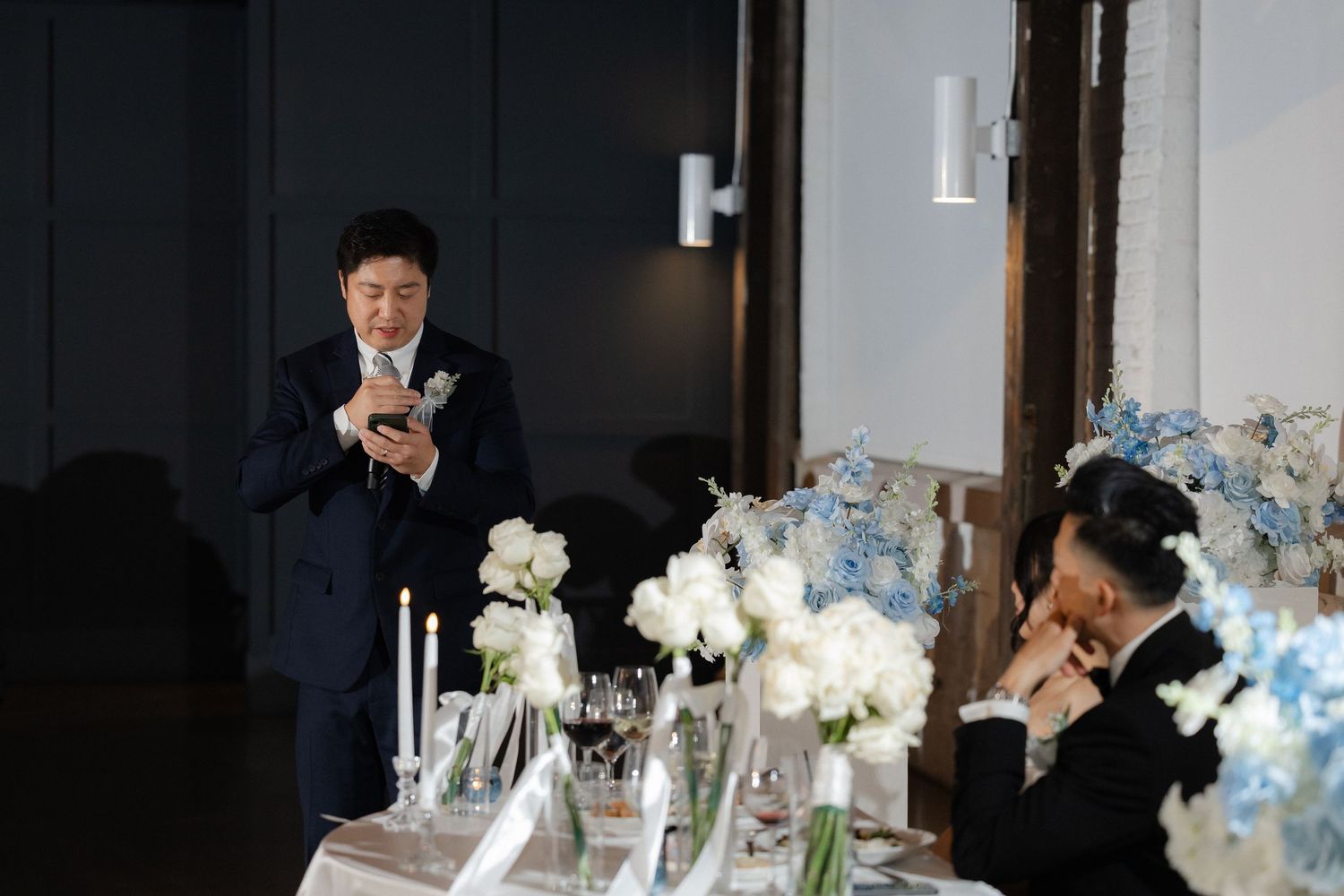Man in suit delivering a heartfelt speech at wedding reception with blue hydrangea floral centerpieces.