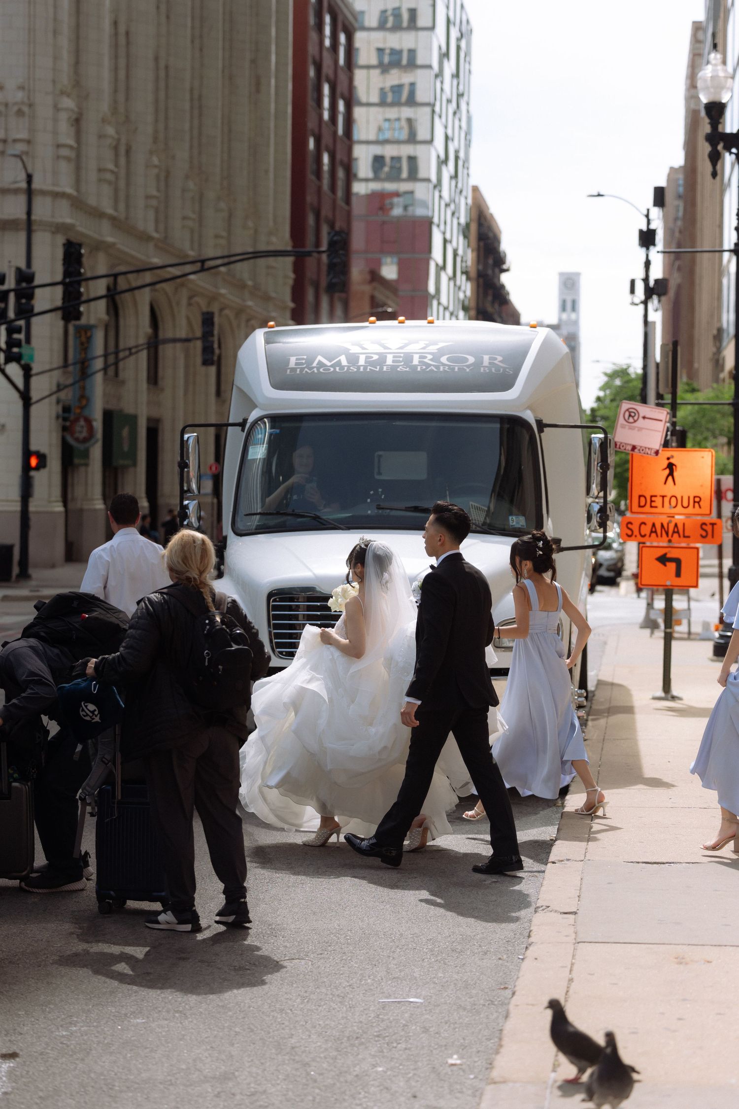 Wedding party walks toward white Emperor limo bus on urban street, bride in white gown with veil.