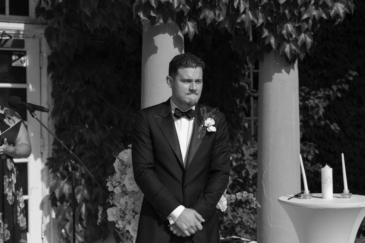 Groom in tuxedo with boutonniere stands at outdoor wedding altar surrounded by ivy-covered columns in black and white photo.