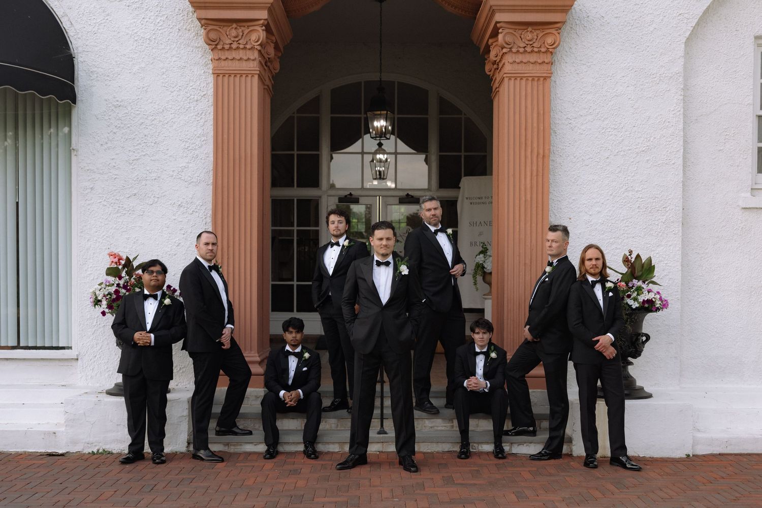 Groom and groomsmen in black tuxedos pose on steps of elegant white building with pink columns at wedding.