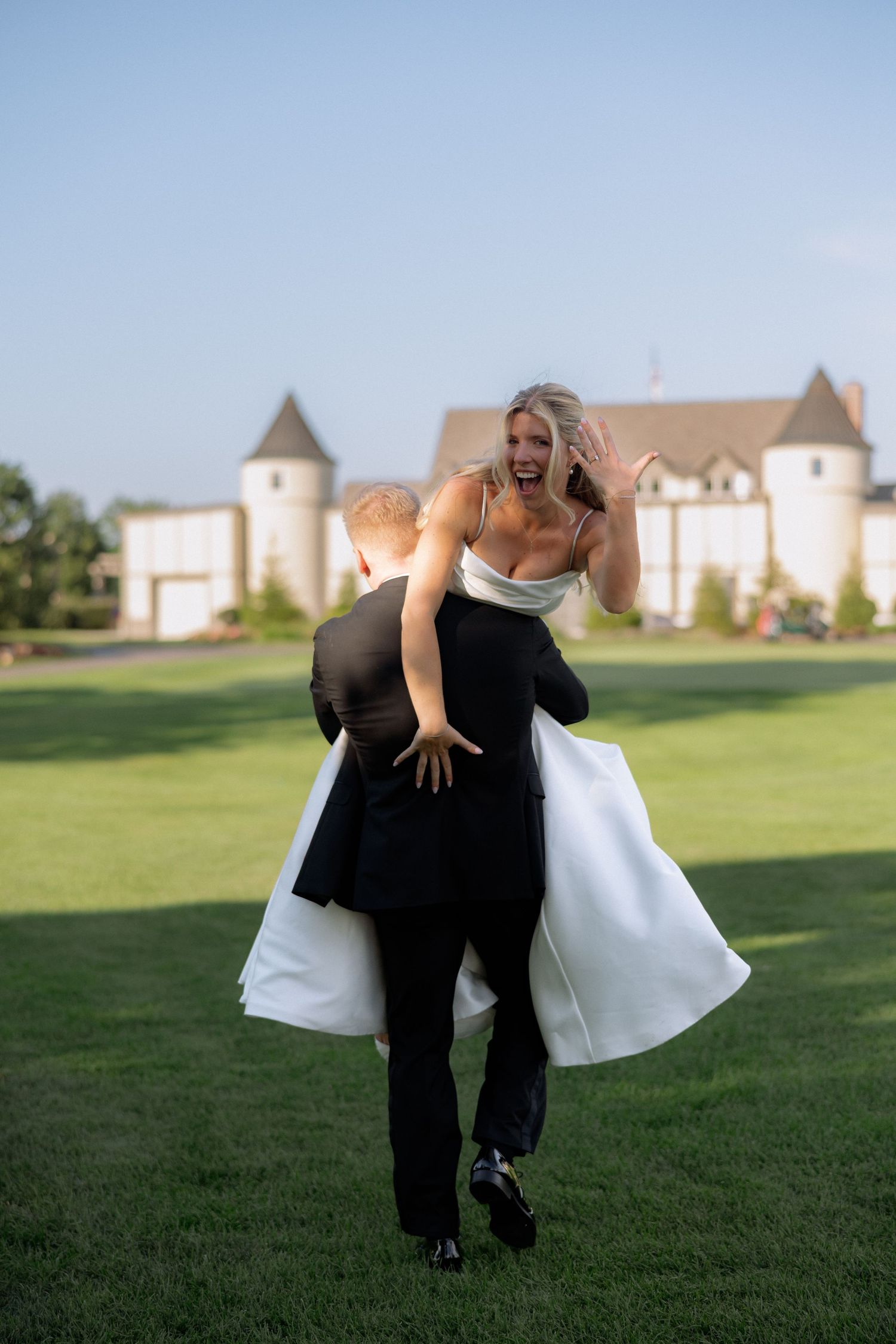 Groom carries laughing bride across green lawn at elegant castle wedding venue.