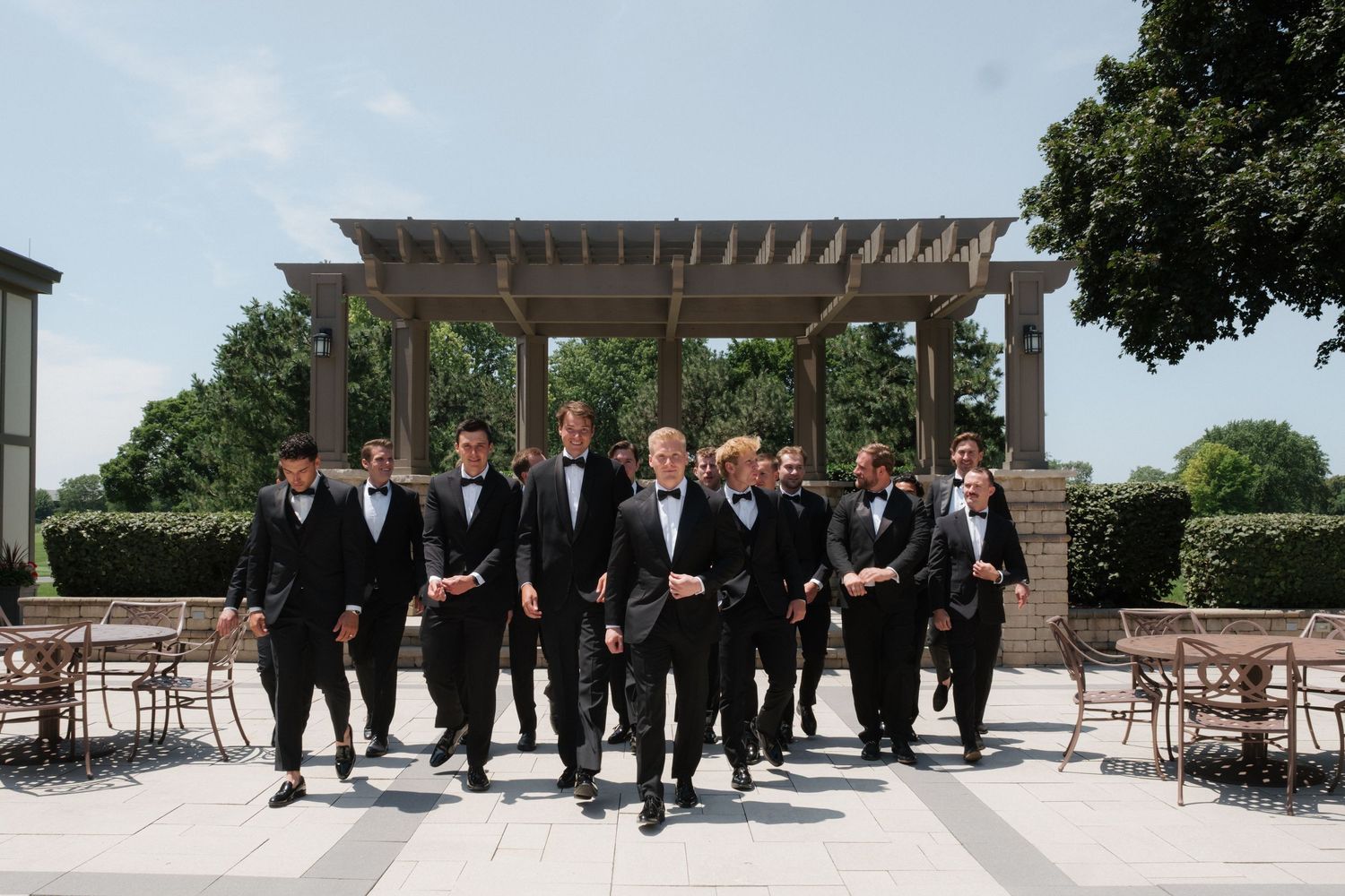 Groom and groomsmen in black tuxedos walking confidently outdoors on a sunny wedding day.
