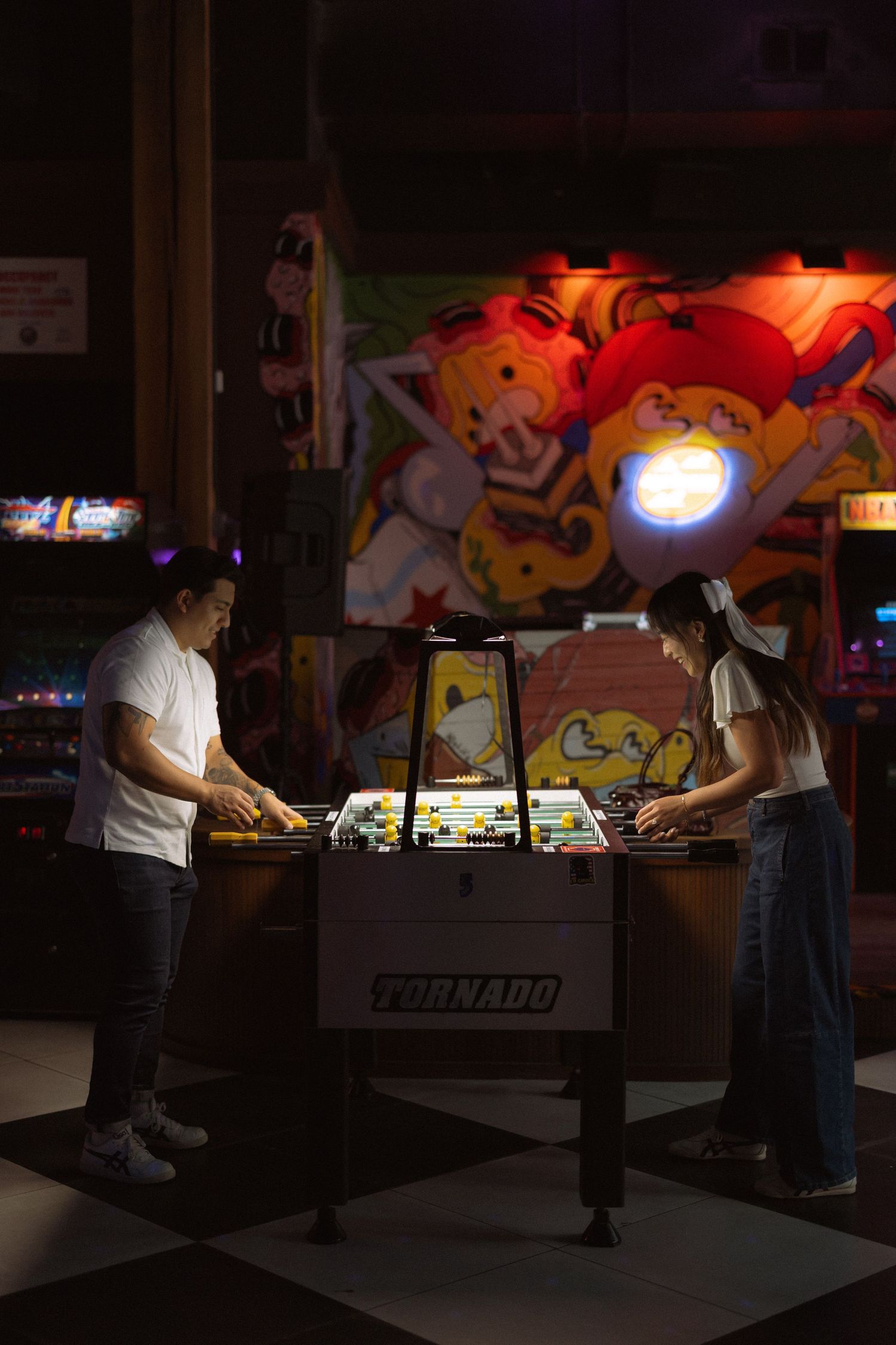 Two people playing foosball on a Tornado table in a dimly lit arcade with colorful game machines in the background.