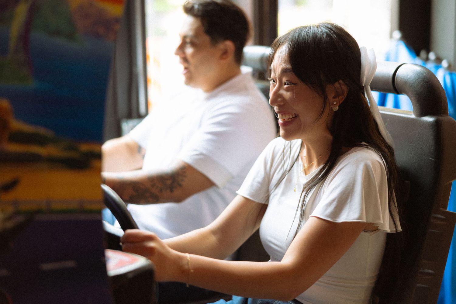 Laughing woman holding a steering wheel at an arcade racing game beside a man.