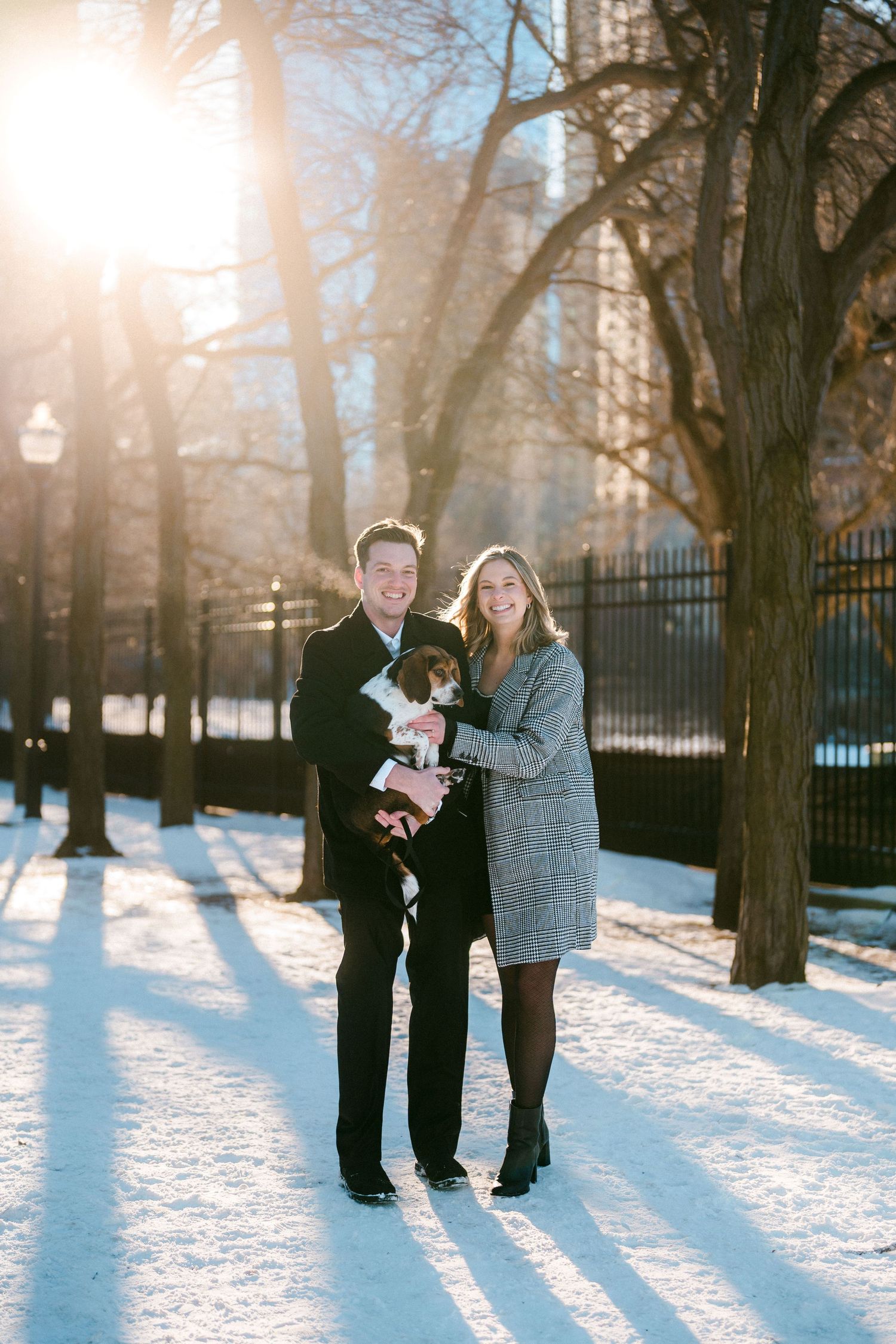 Smiling couple holding their dressed-up beagle in a sunny snow-covered park setting.