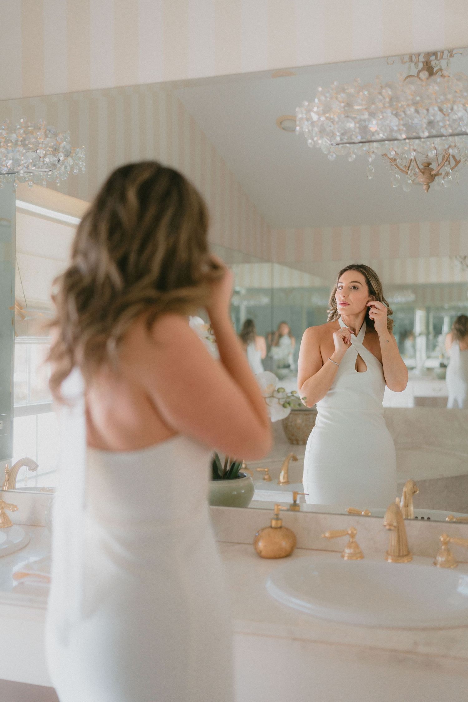 Bride in white halter dress putting on earrings while looking in elegant bathroom mirror.