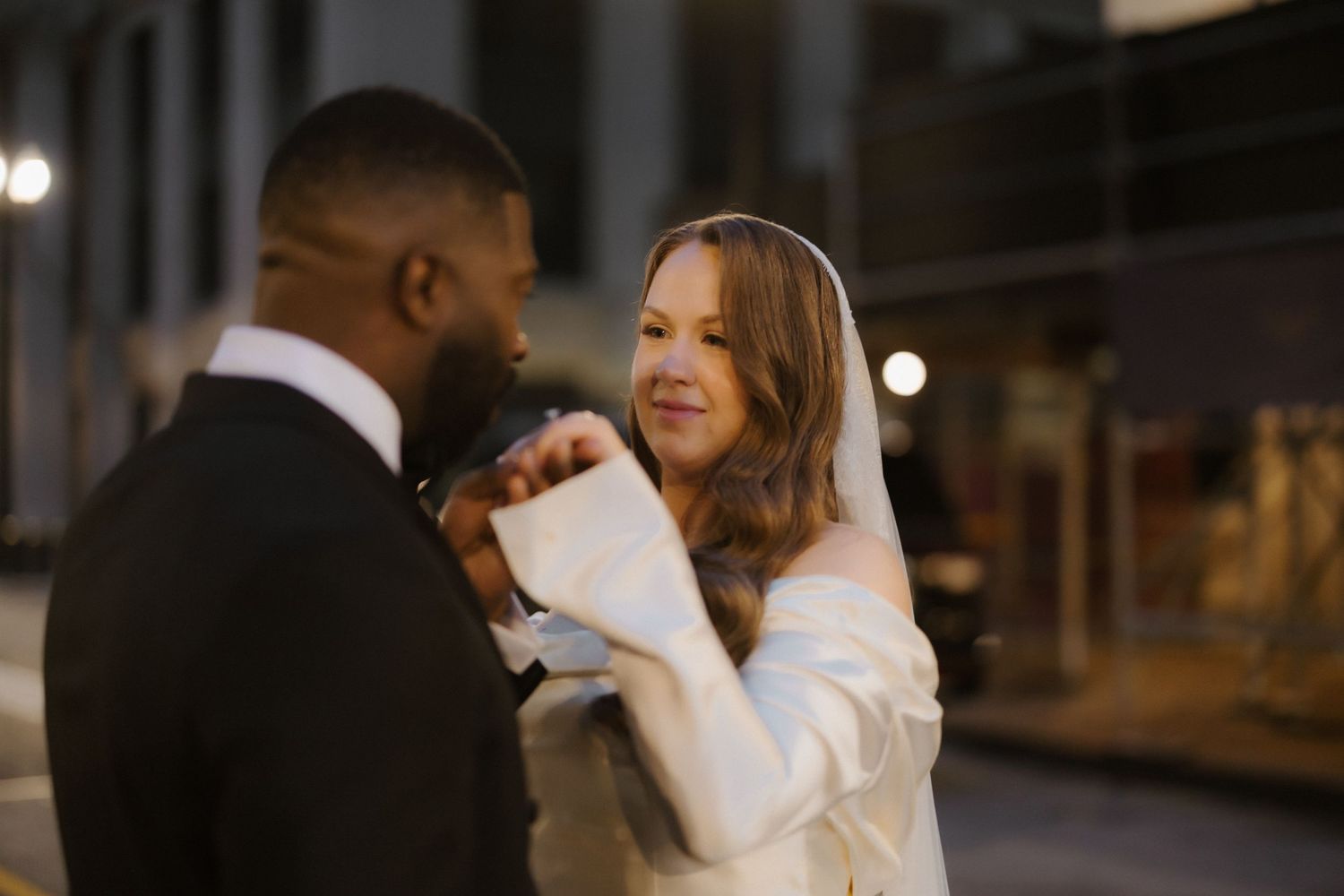 Interracial couple sharing a romantic moment outdoors at night, bride in white off-shoulder gown holding groom's hands.