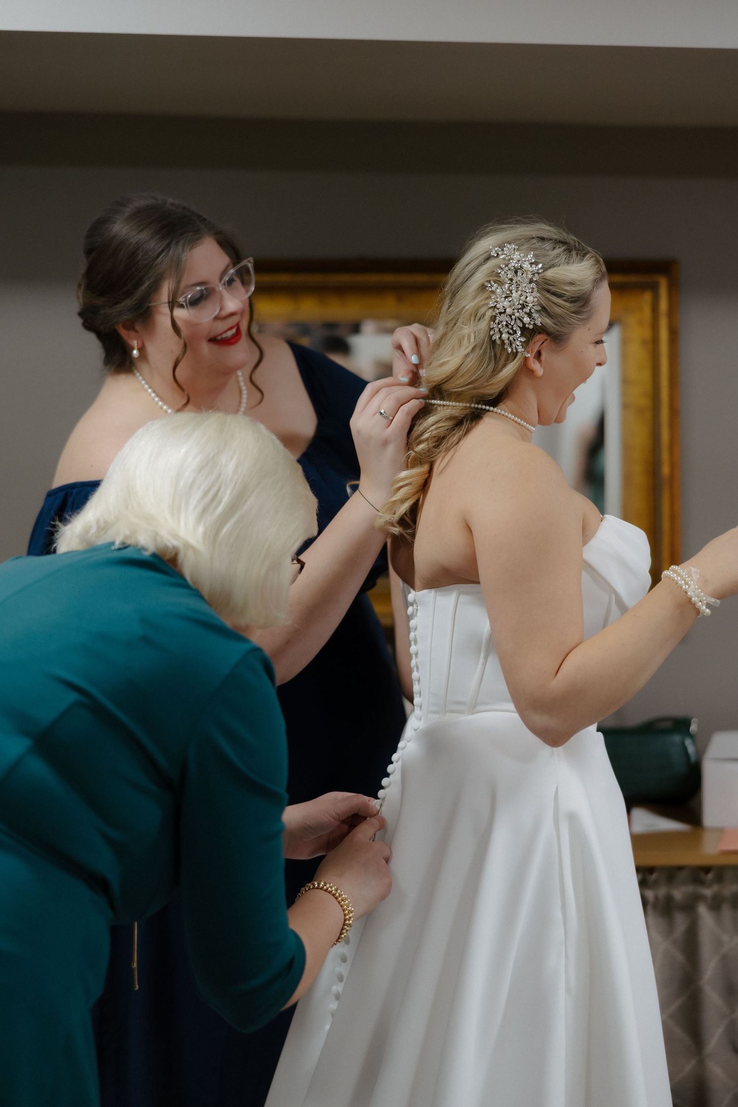 Two women help a bride in a white gown with her hair and dress buttons before her wedding ceremony.