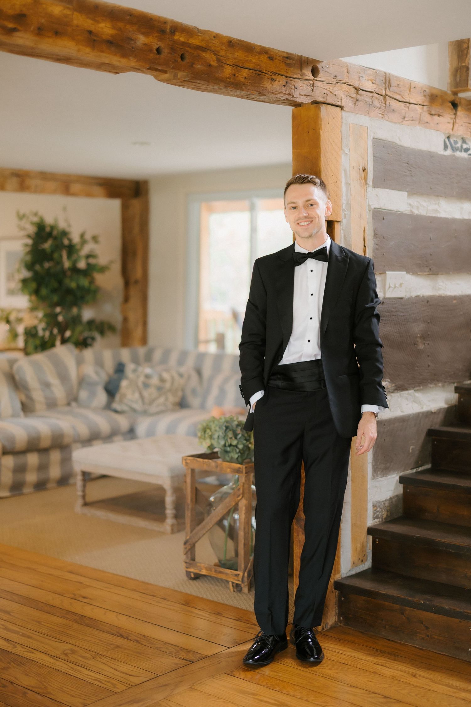 Groom in black tuxedo smiling in rustic log cabin interior with wooden beams and staircase.