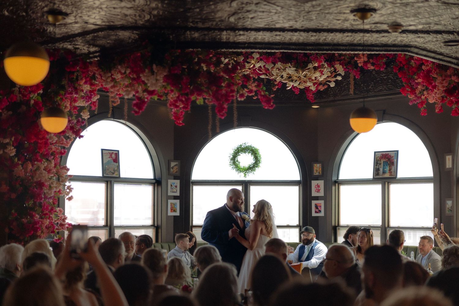 Couple shares first dance surrounded by guests in floral-decorated venue with arched windows and wreath.