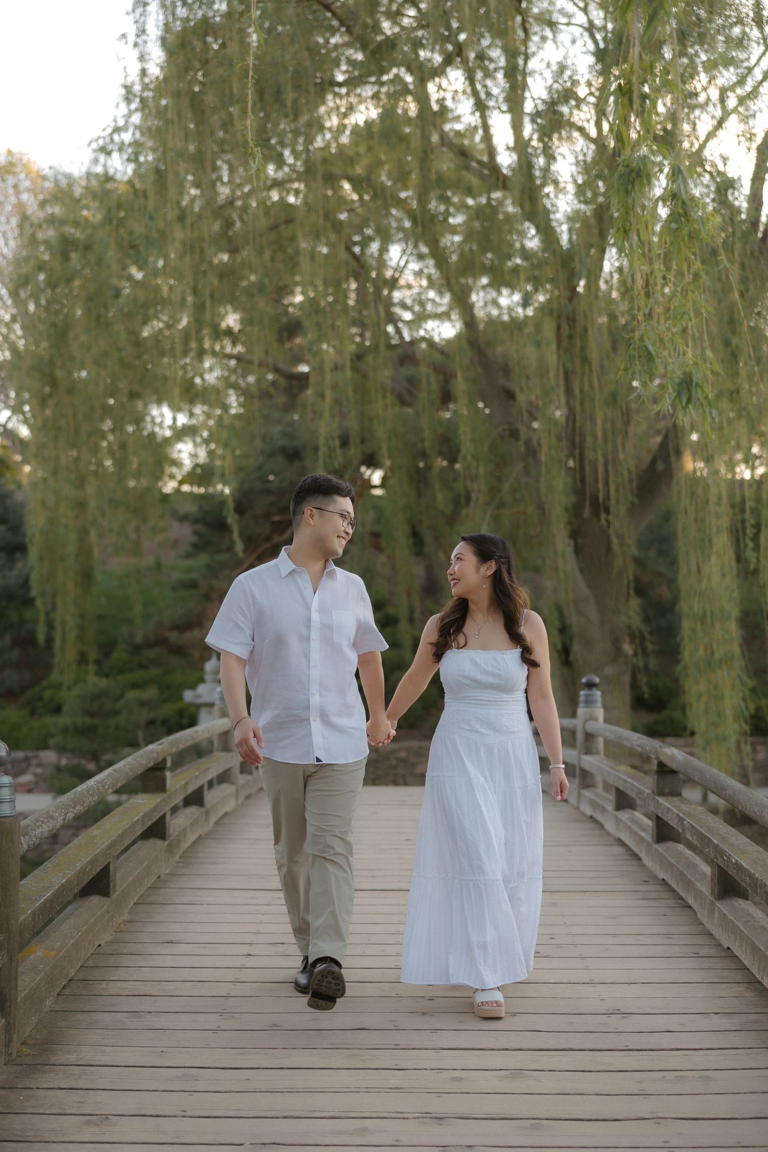 Couple in white outfits holding hands walking on wooden bridge surrounded by weeping willow trees.