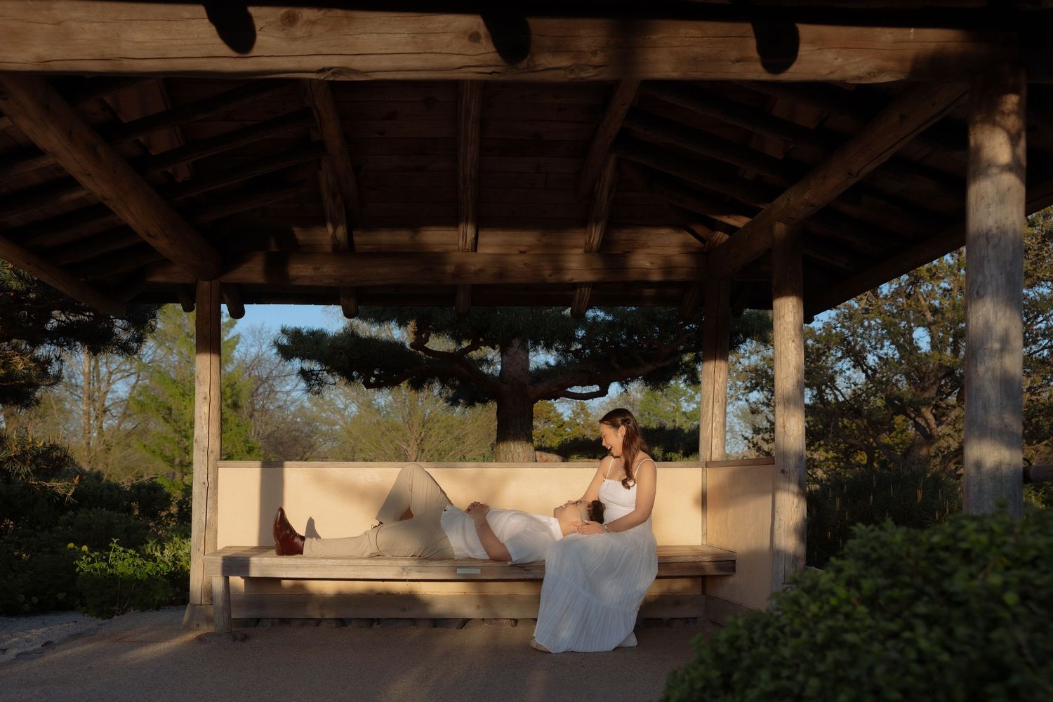 Woman in white dress relaxing on a bench under a wooden pergola surrounded by lush greenery at golden hour.