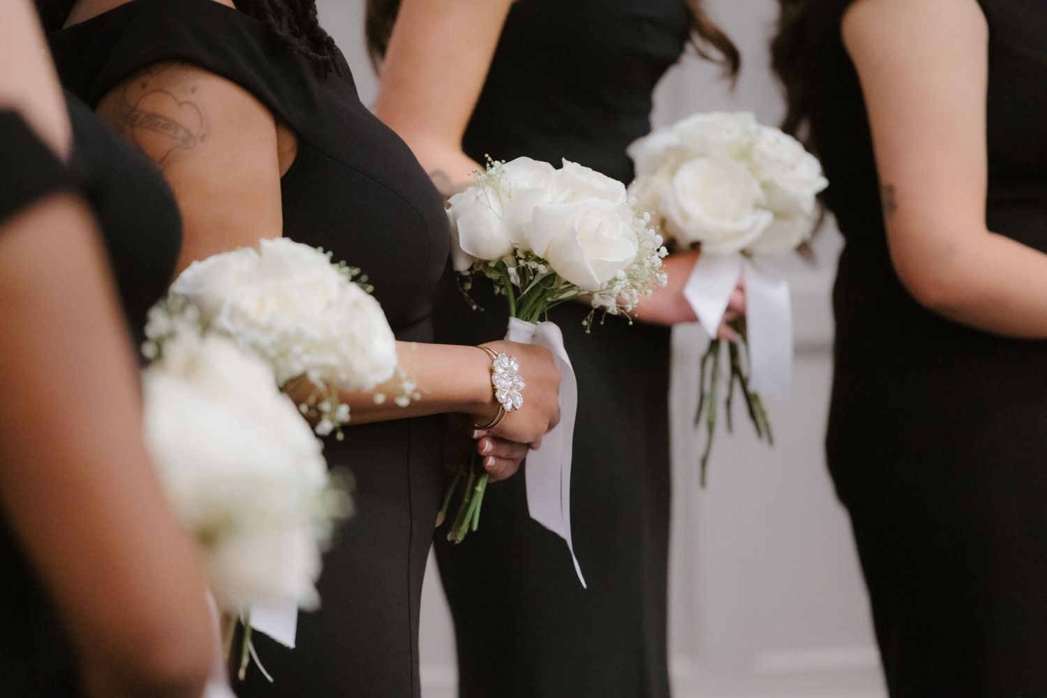 Bridesmaids in black dresses holding white peony bouquets with ribbon handles at a wedding ceremony.