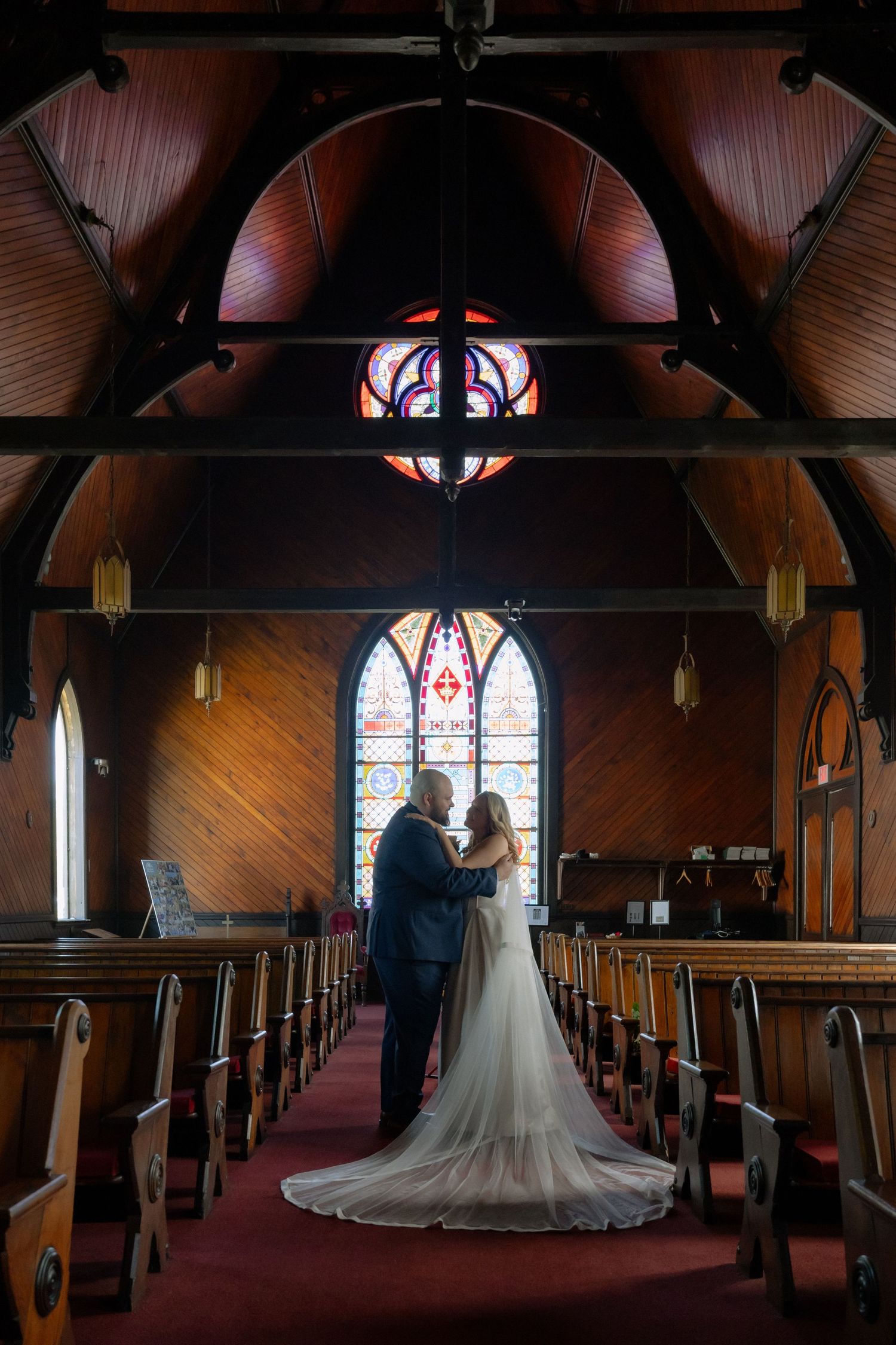 Bride and groom embrace at the altar of a stunning wooden chapel with colorful stained glass windows.