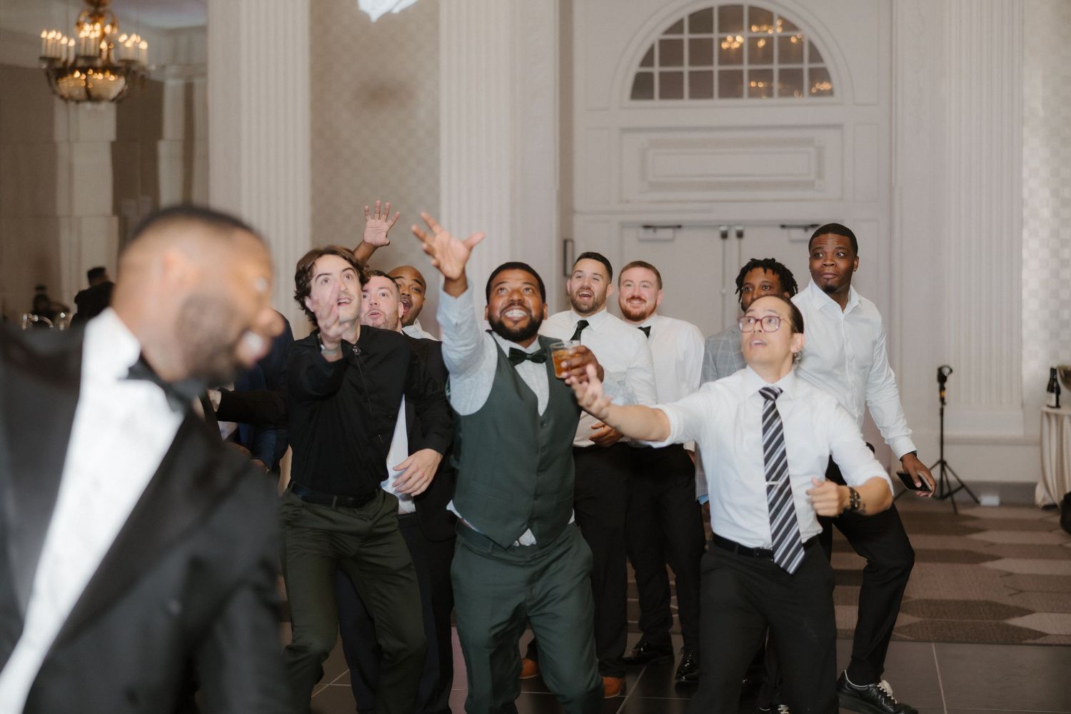 Groomsmen in suits laugh and dance together in an elegant ballroom during a lively wedding reception celebration.