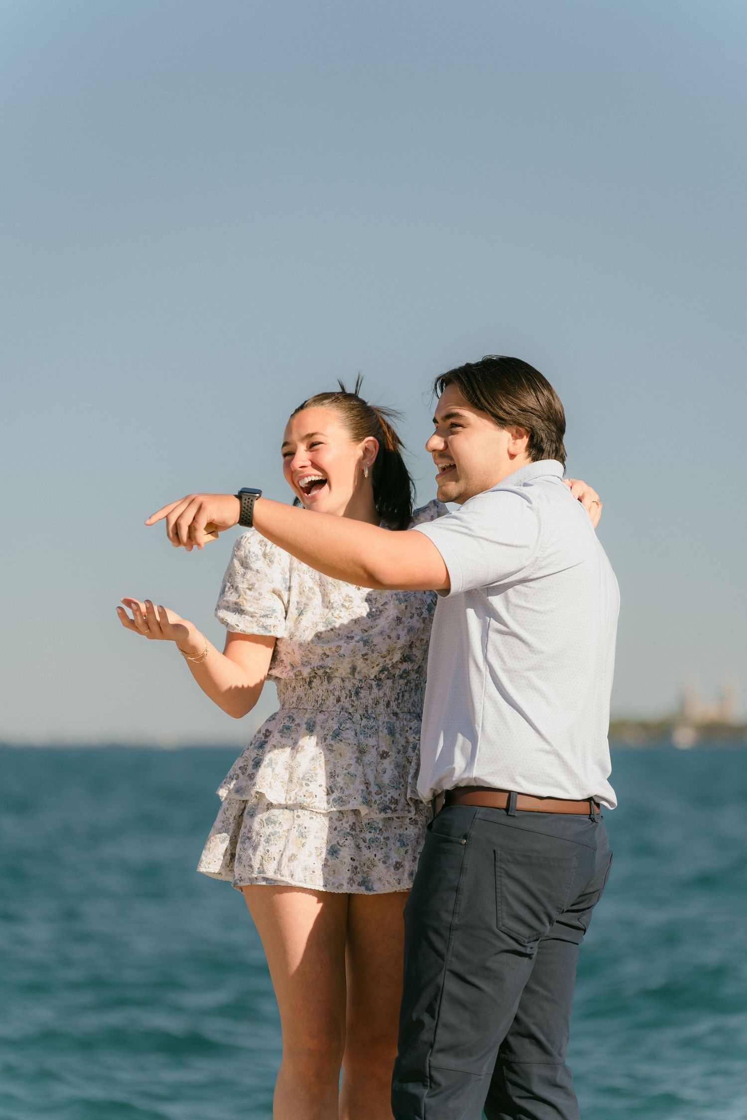 Joyful woman showing engagement ring while embracing partner by the lake.