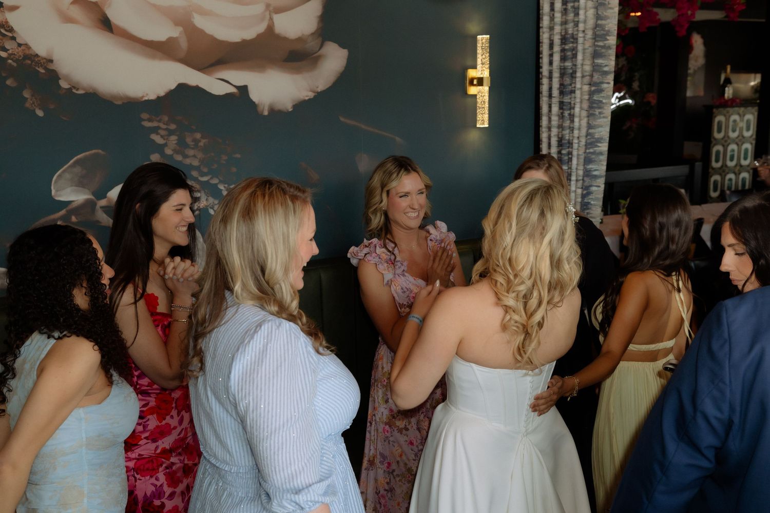 Bride in white gown surrounded by guests at wedding reception near teal floral wall.