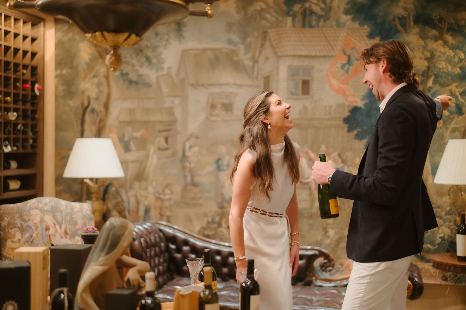 Laughing couple opening champagne bottle in ornate room with tapestry wall decor.