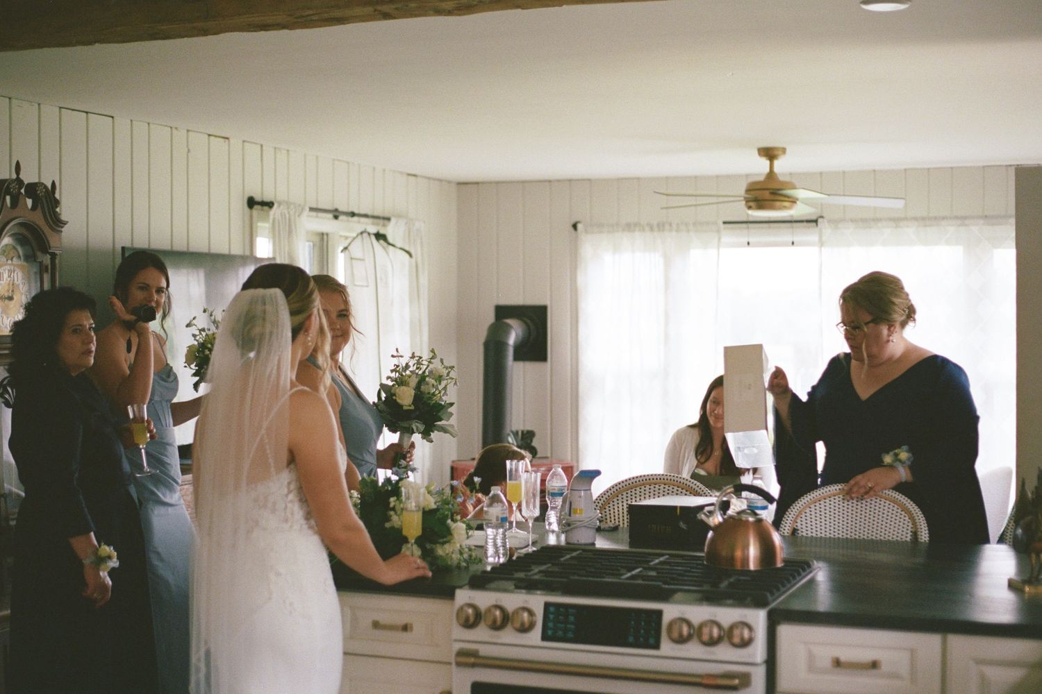 Bride in white gown and veil stands in kitchen surrounded by wedding party during bridal preparations.
