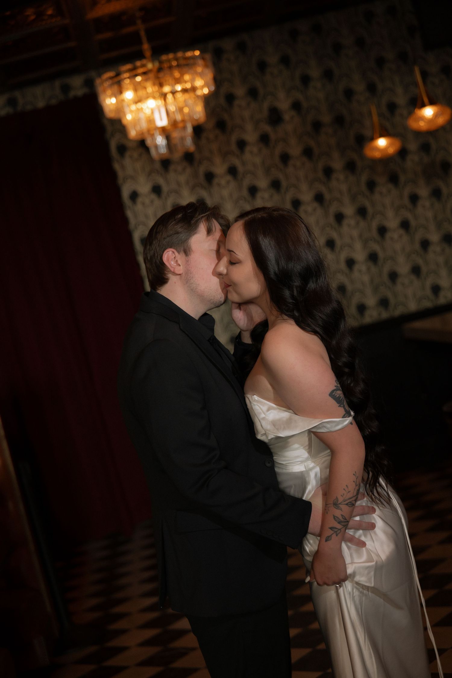 A couple shares a romantic kiss in an elegant venue with chandelier lighting and patterned wallpaper.