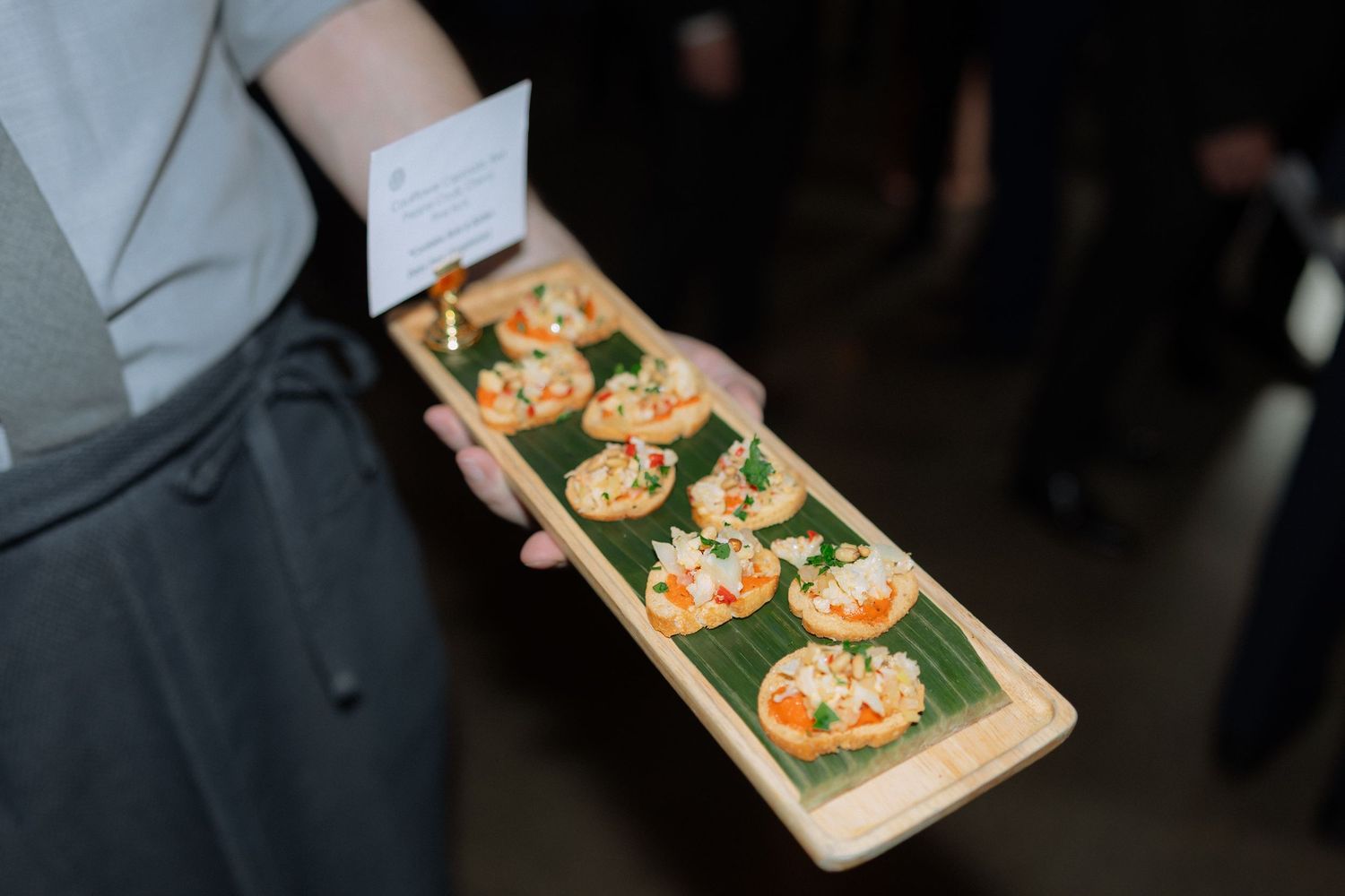 Chef holding a wooden platter of elegant appetizer canapés garnished with vegetables on banana leaf at catered event.