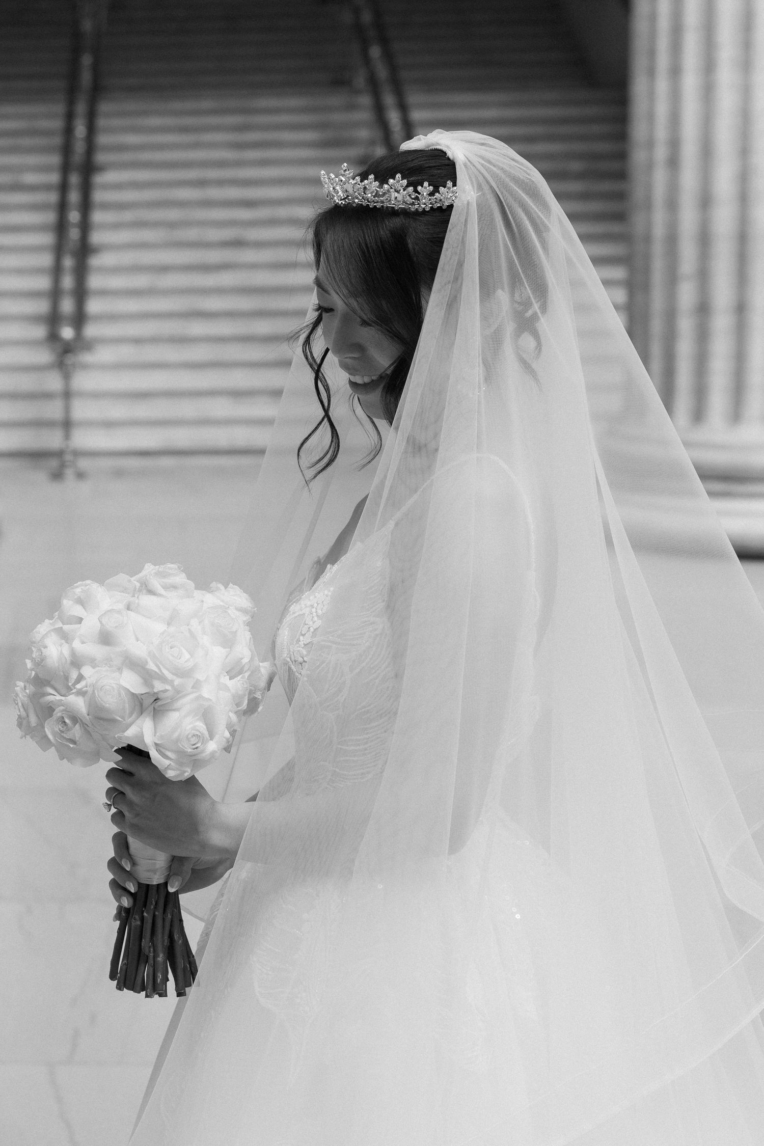 Black and white portrait of bride in tiara and veil gazing down at her white rose bouquet.