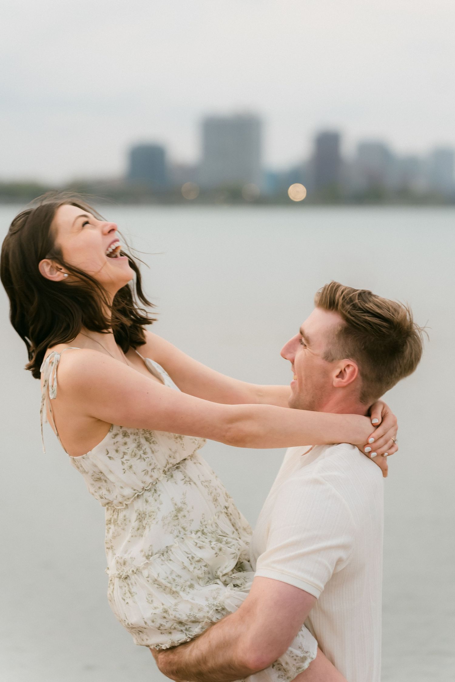 A joyful couple laughing together by the waterfront, man lifting woman in a floral dress with city skyline behind.