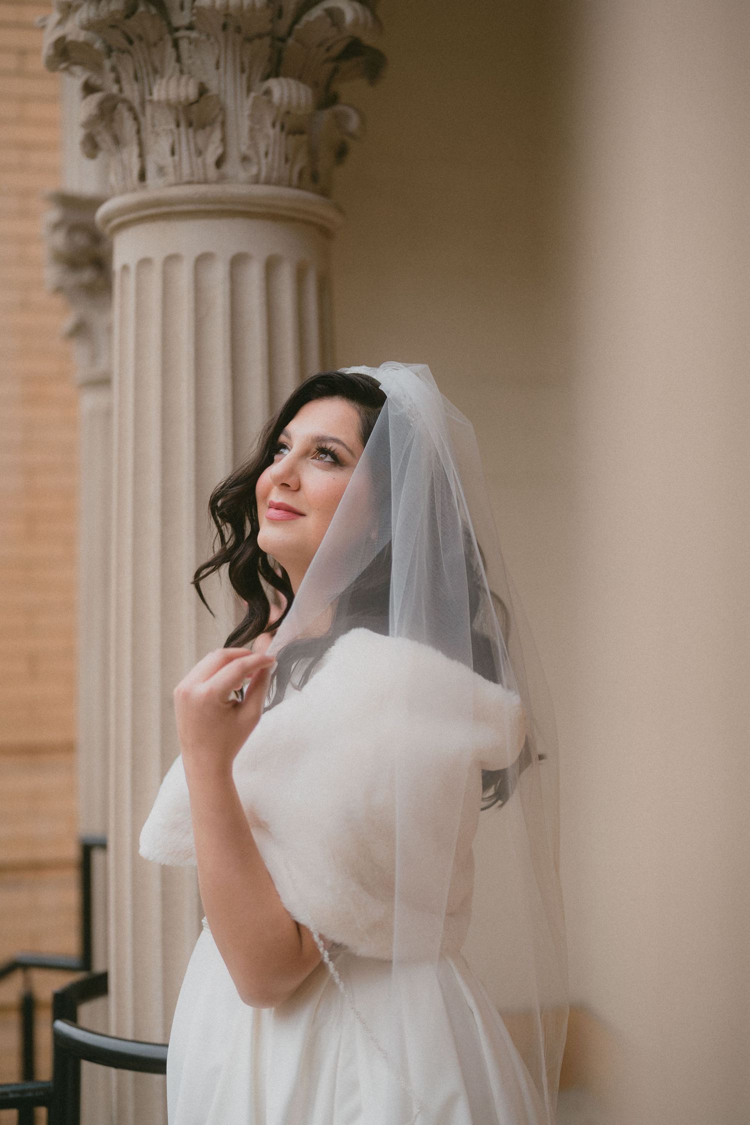 Beautiful bride in white off-shoulder gown and veil poses beside ornate classical column before wedding ceremony.
