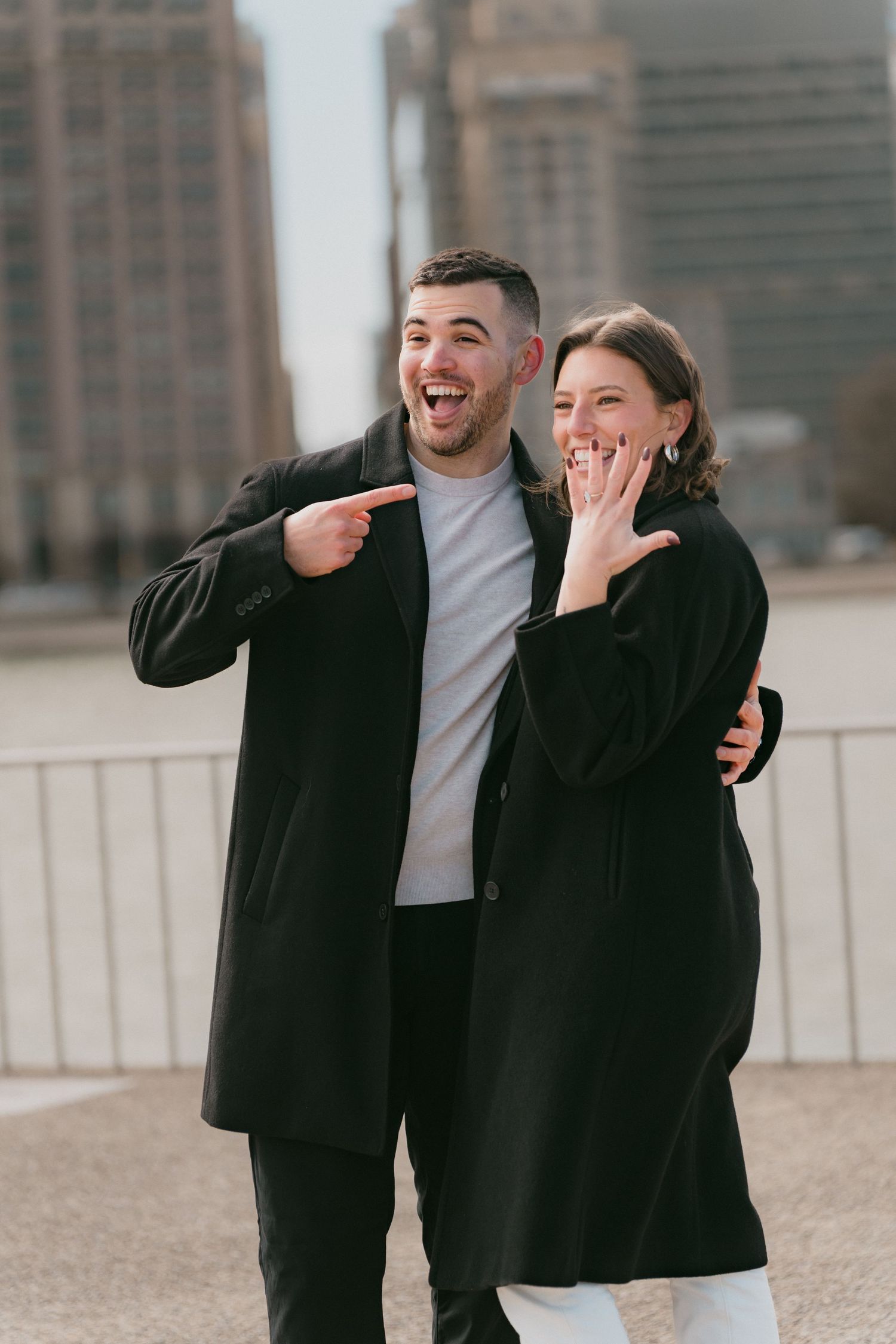 Happy newly engaged couple posing in Chicago, man pointing at partners engagement ring as she shows her hand.