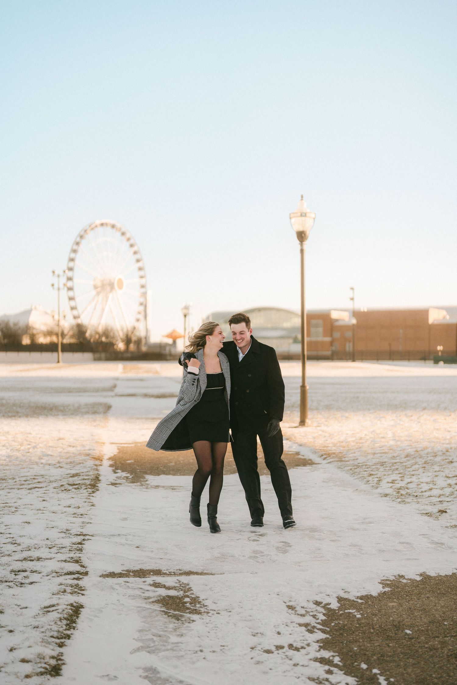 Couple walking together in snow near Ferris wheel under clear winter sky.