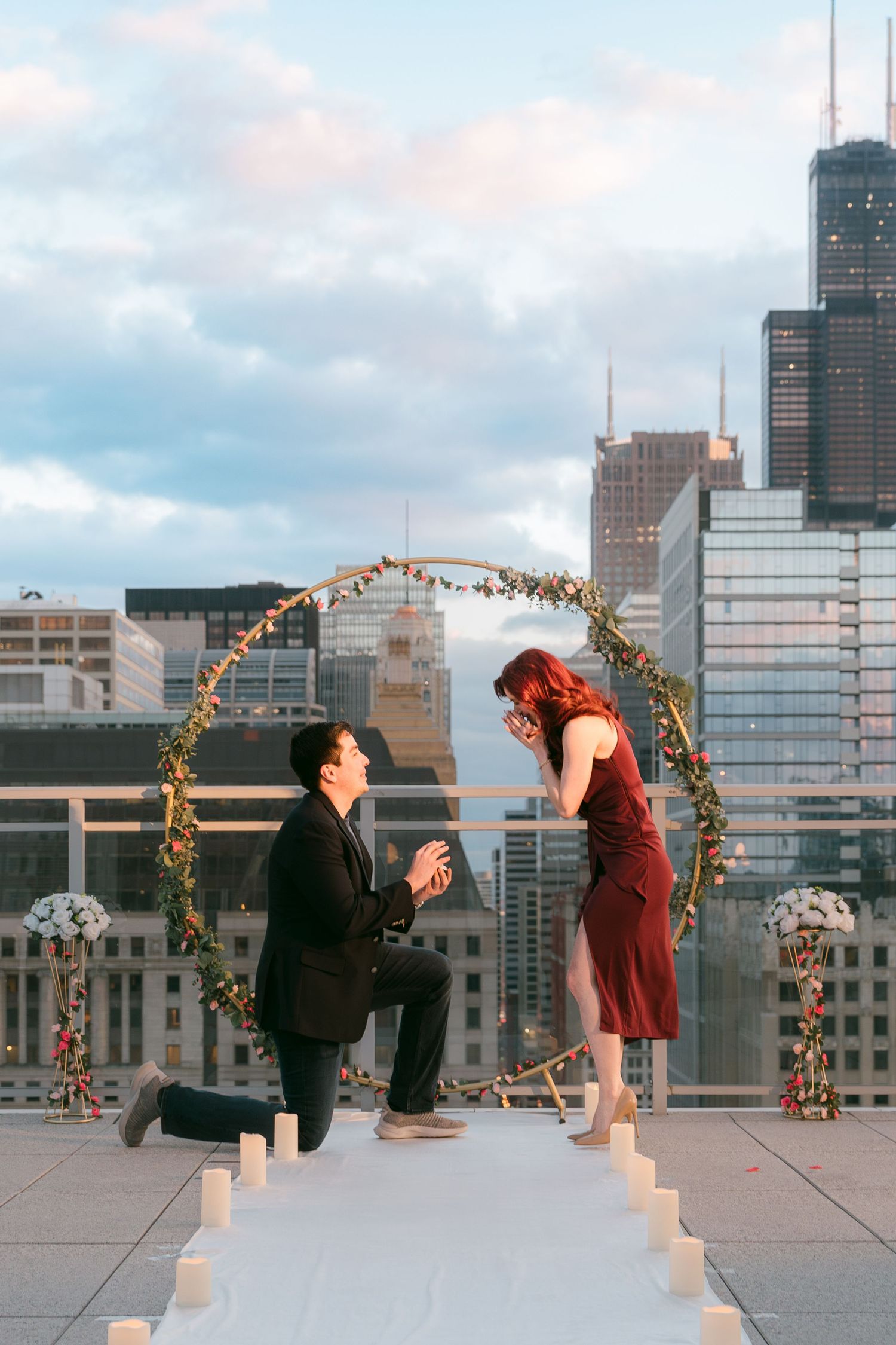 Man on one knee proposing to woman in burgundy dress under floral arch on Chicago rooftop.