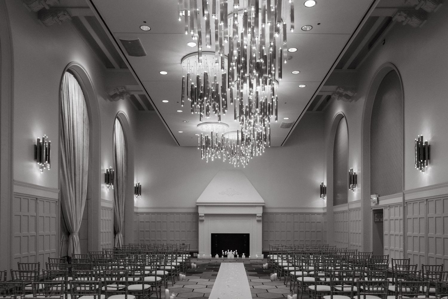 Elegant ballroom with grand crystal chandelier, arched windows, fireplace, and rows of chairs set for a formal wedding ceremony.