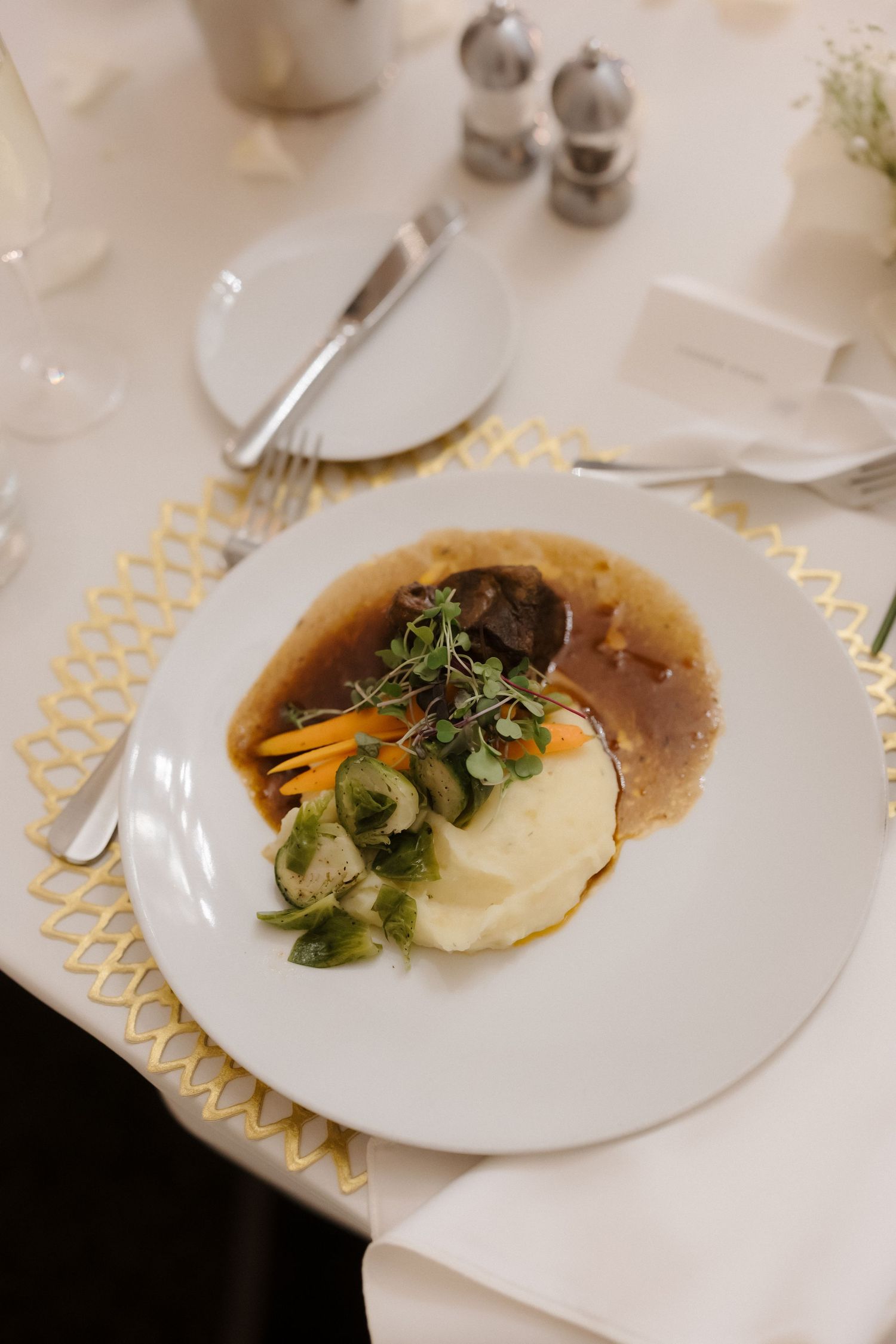 Elegant plated dinner with mashed potatoes, braised meat, carrots, Brussels sprouts, and rich gravy garnished with microgreens.