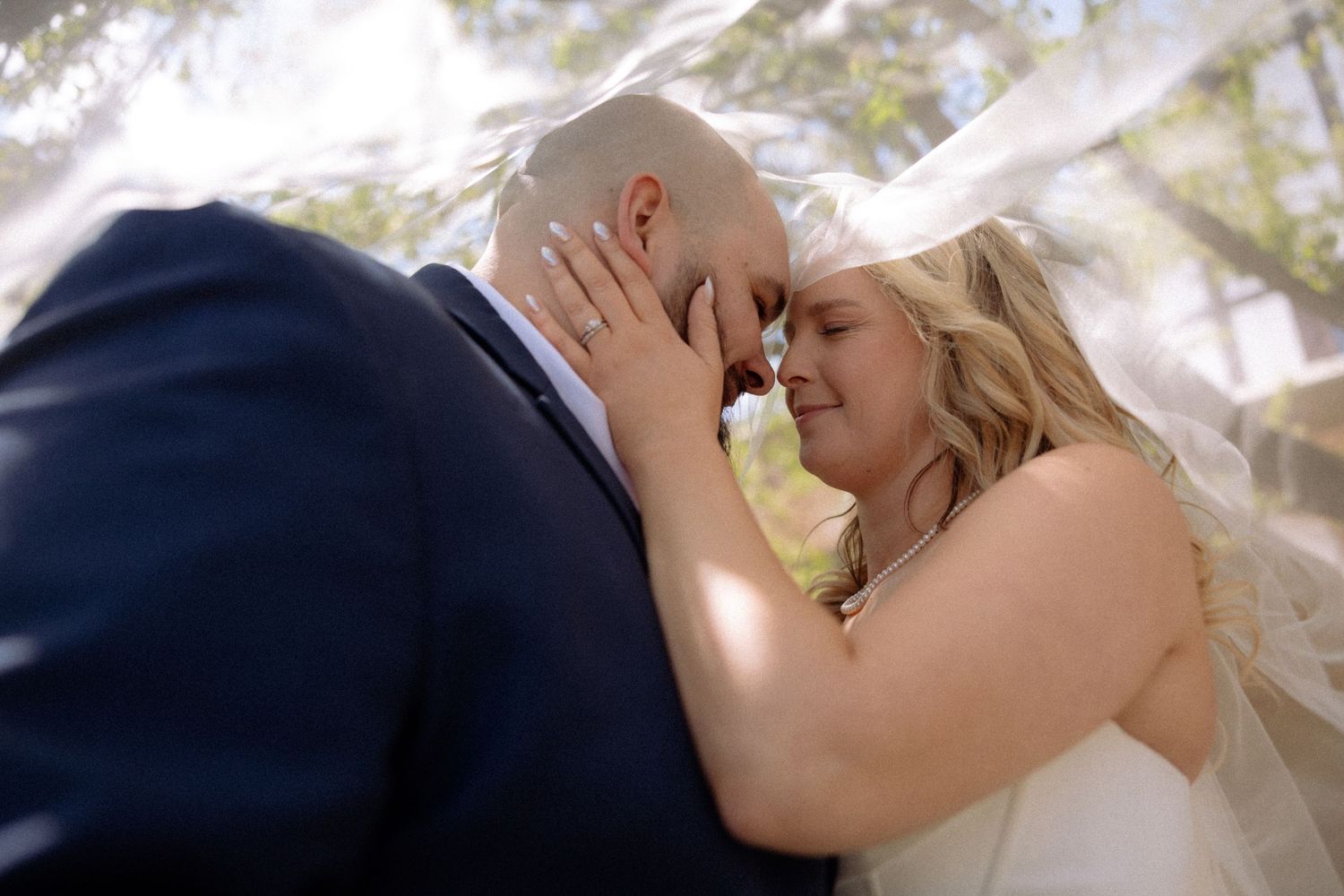 A bride and groom share an intimate forehead touch under a flowing white veil outdoors on their wedding day.