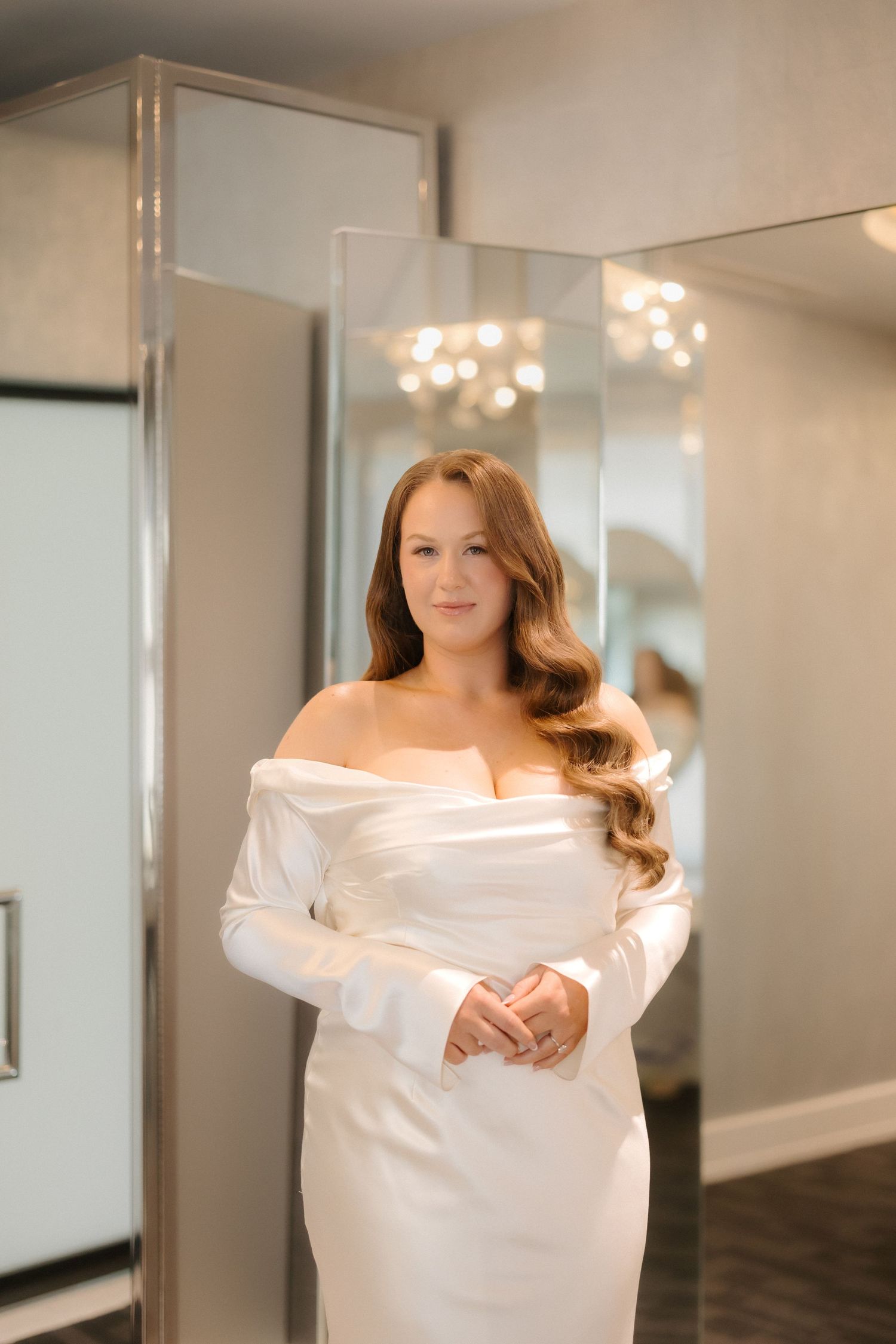 Bride in elegant off-shoulder white satin gown with long sleeves poses in front of mirrors in a bridal boutique.