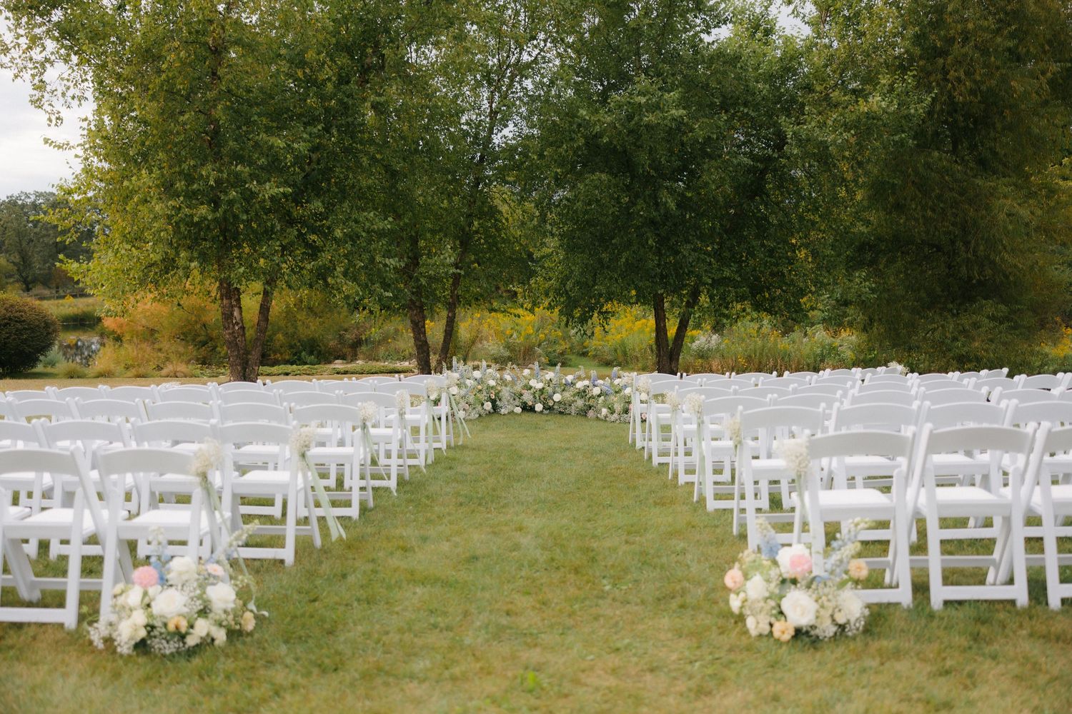 Elegant outdoor wedding ceremony setup with white chairs and floral arrangements on lush green lawn.