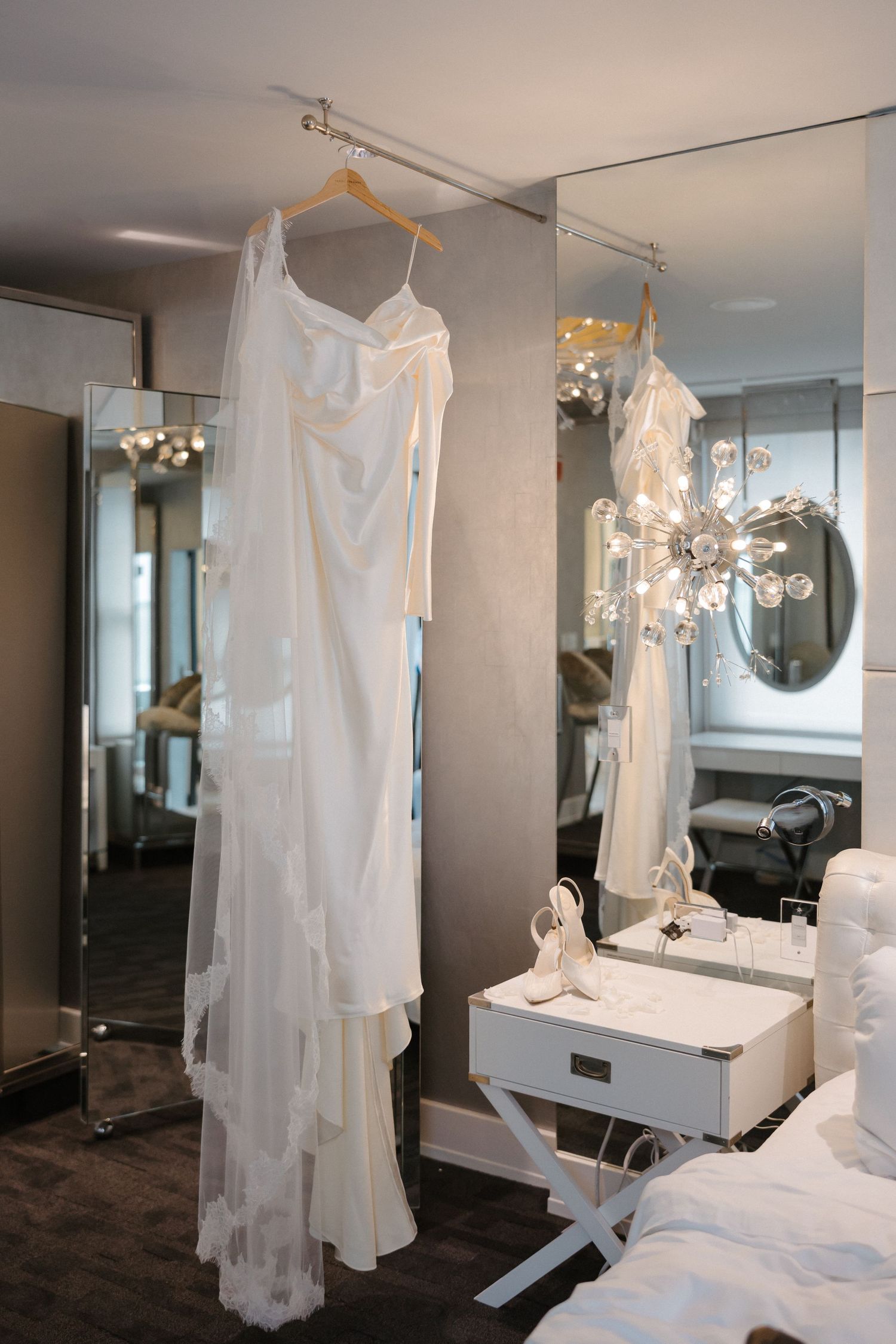 Elegant white wedding dress hanging on a hanger beside a mirror and vanity table in a bridal suite.