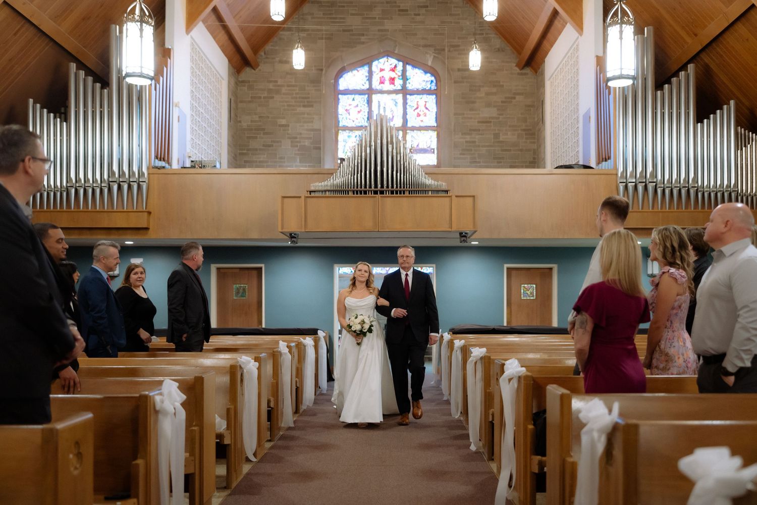 Bride walks down church aisle with escort past wooden pews, pipe organ visible in background during wedding ceremony.