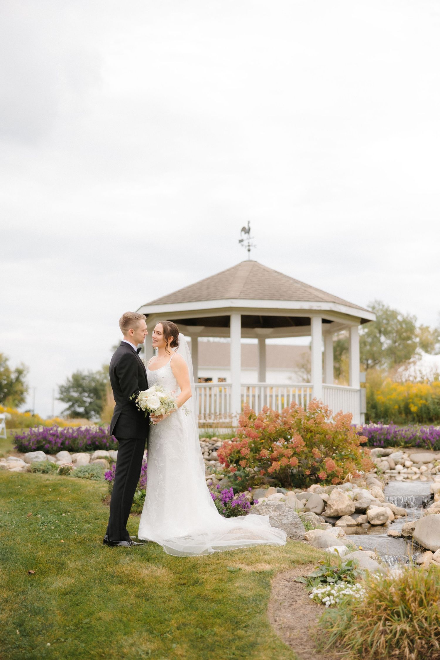 Bride and groom posing near a white gazebo surrounded by colorful garden flowers.