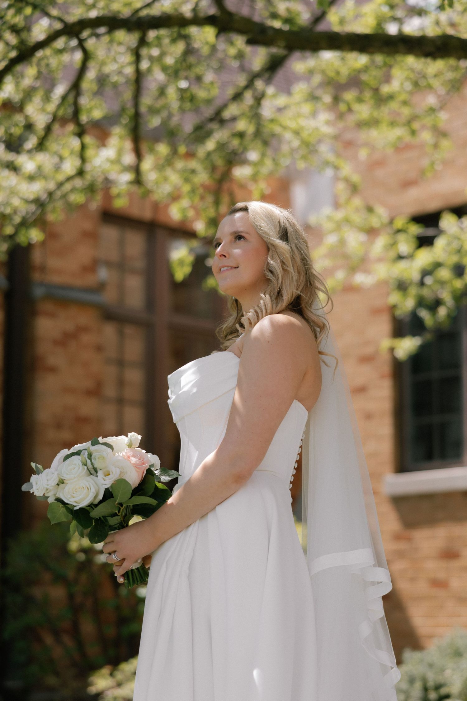 Bride in white strapless gown holding floral bouquet, standing outdoors near brick building with blooming trees.