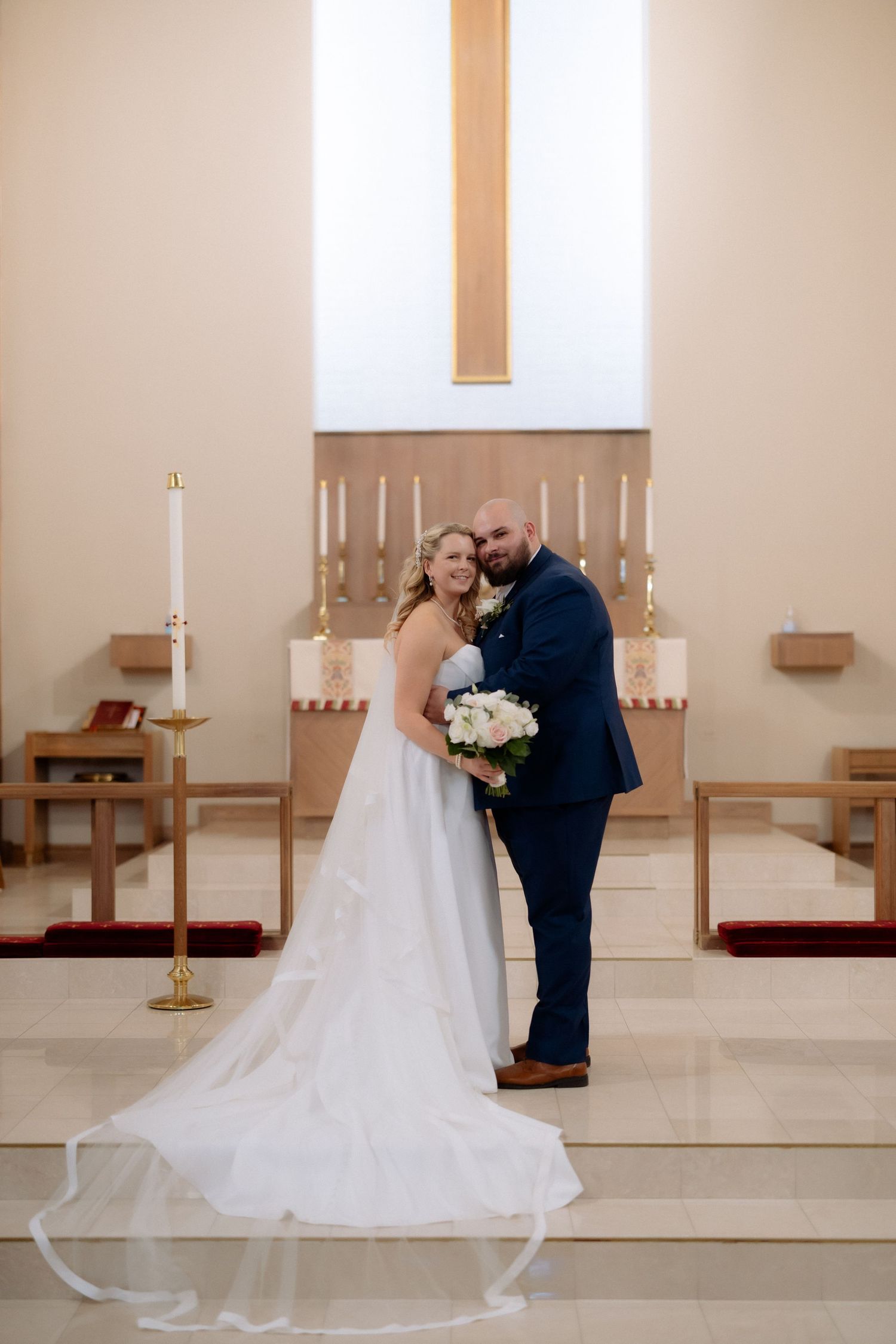 Bride and groom pose together at church altar, bride in white gown with veil, groom in navy suit holding bouquet.