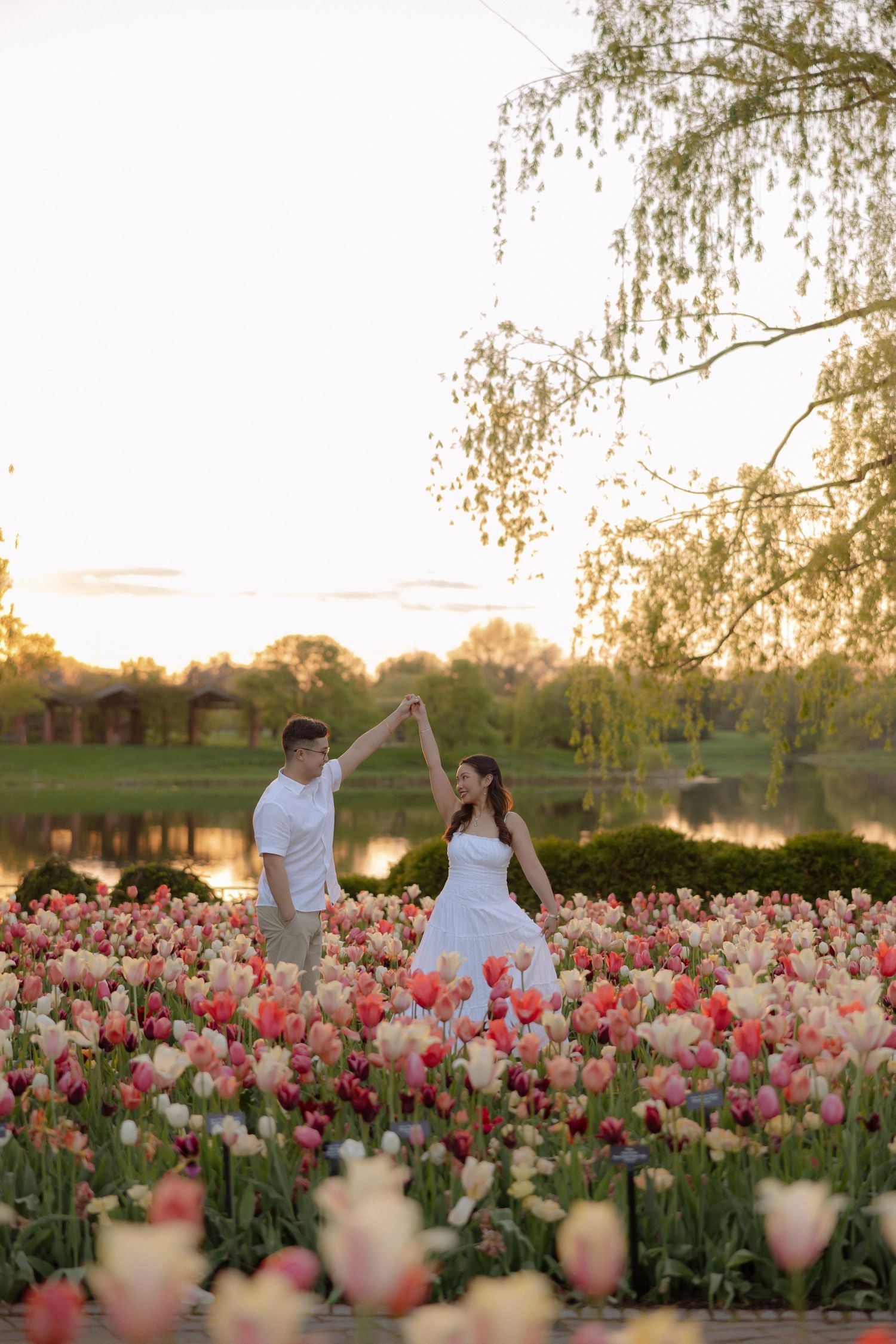 Couple dancing in a vibrant tulip field at sunset near a serene lake, surrounded by pink and white blooms.