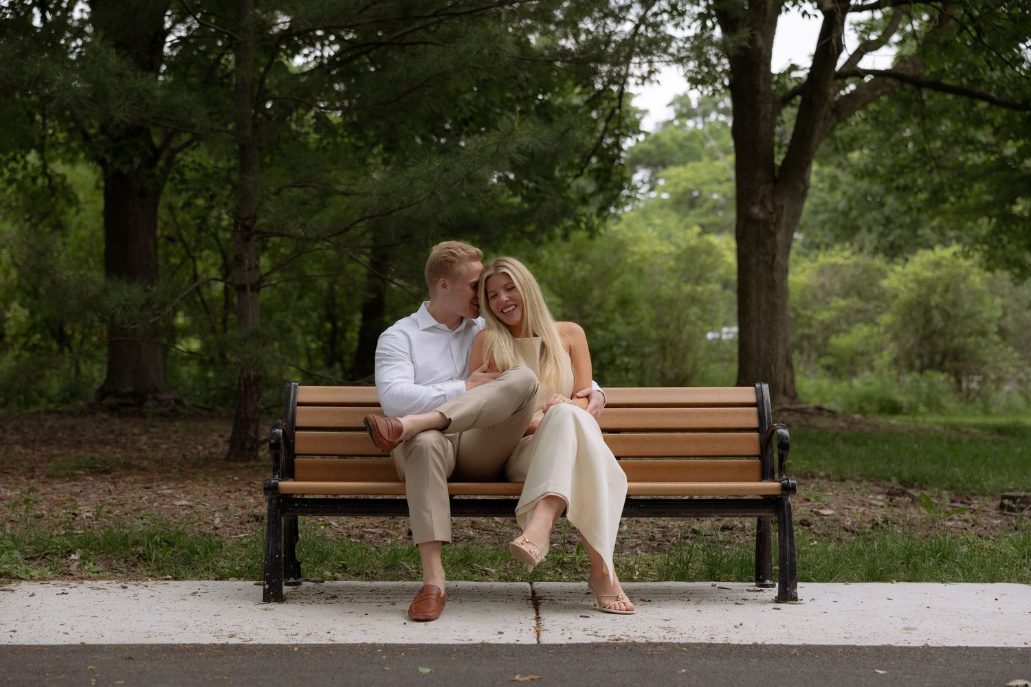 Couple sitting close together on a wooden park bench surrounded by lush green trees on a sunny day.