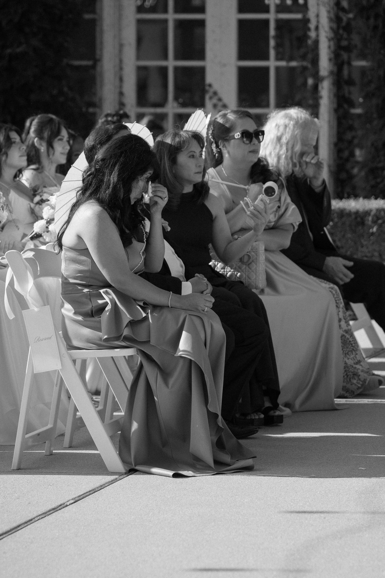 Black and white photo of emotional wedding guests seated in white chairs during outdoor ceremony.